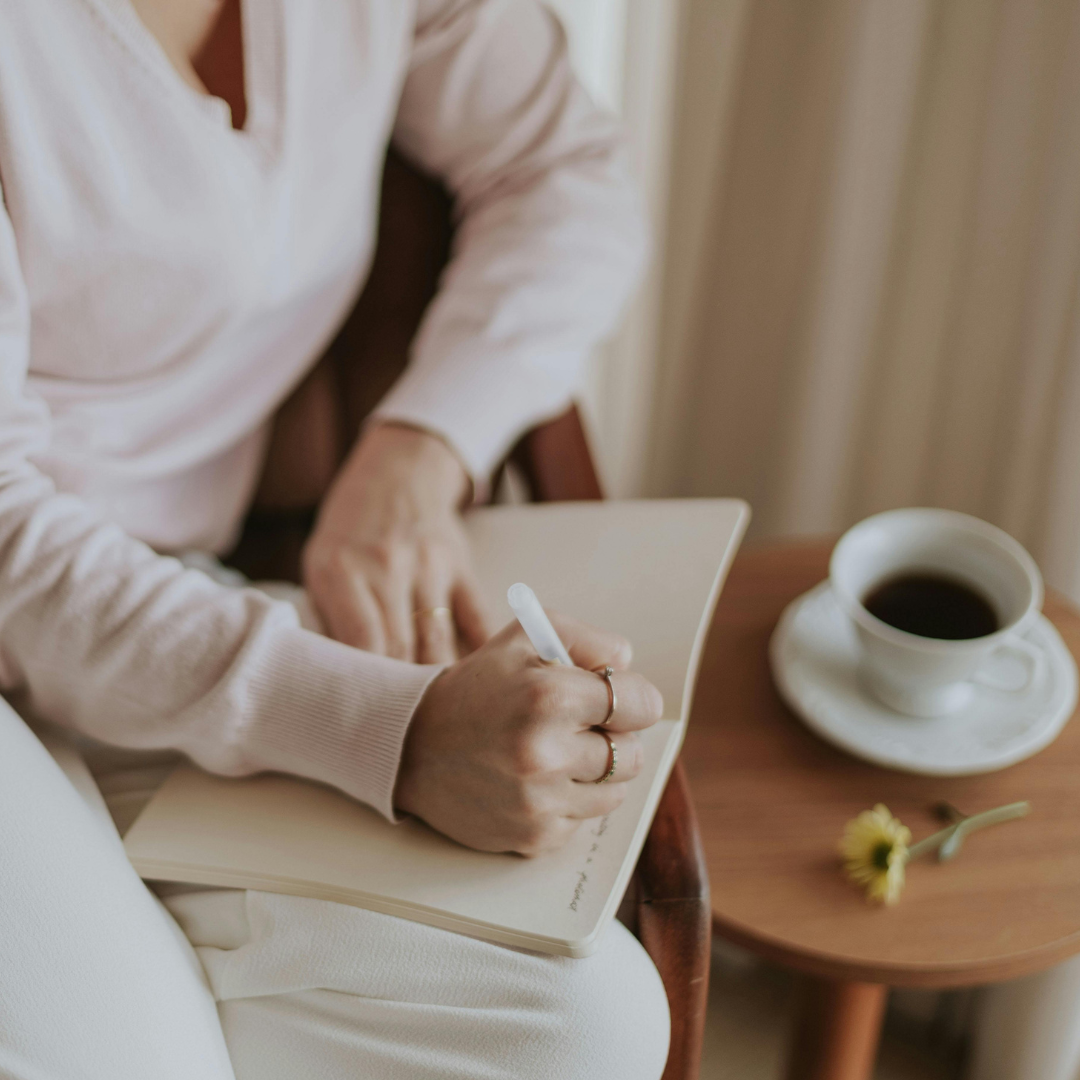 Person writing in a notebook with a cup of coffee on a wooden table nearby.