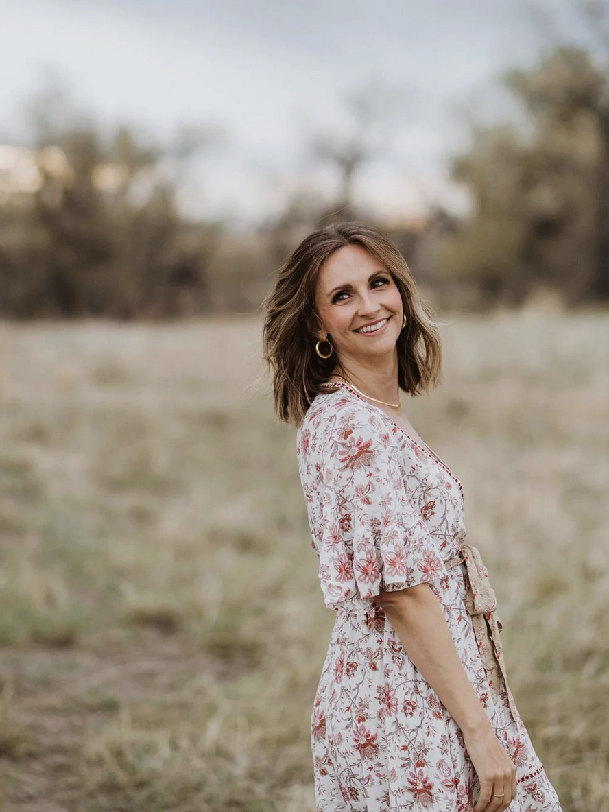 A woman with shoulder-length brown hair, wearing a white dress with pink floral patterns, standing outdoors in a natural setting with blurred trees in the background, smiling and looking slightly to her left.