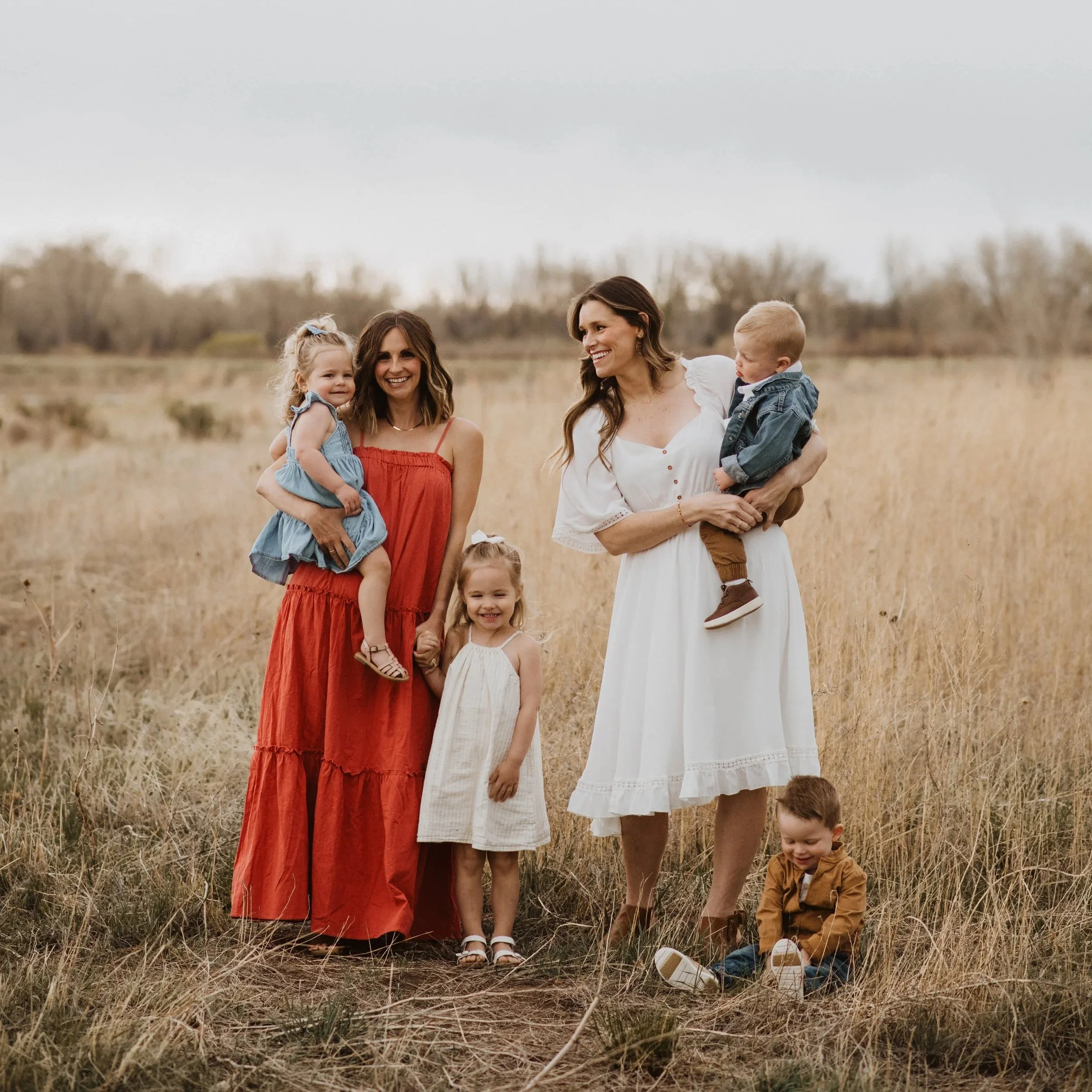 Group of women and children standing in a grassy field, smiling and holding children, with trees in the background.