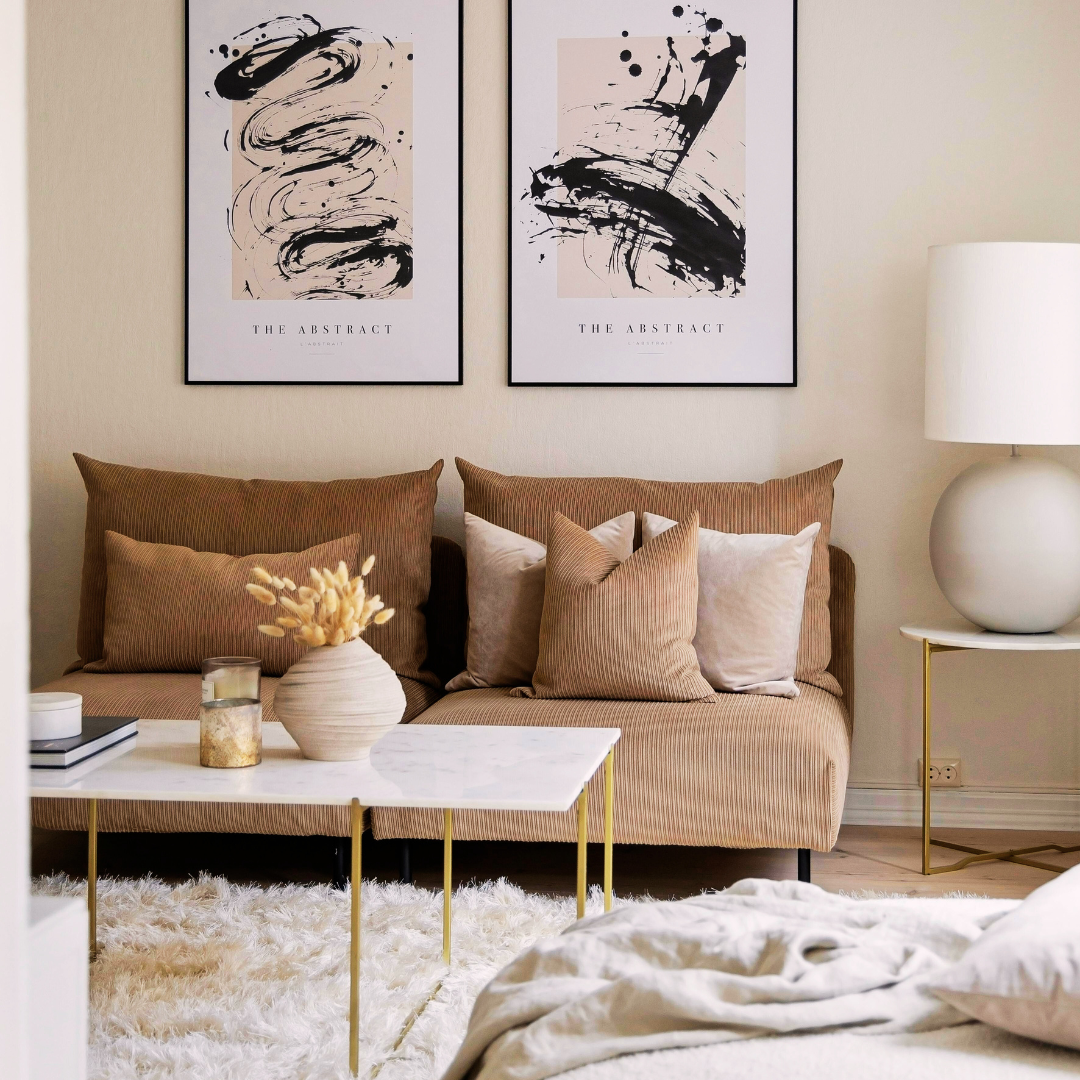 A living room with a beige couch and pillows, a marble coffee table with decorative items, two black and white abstract art prints on the wall, and a white lamp on a side table.