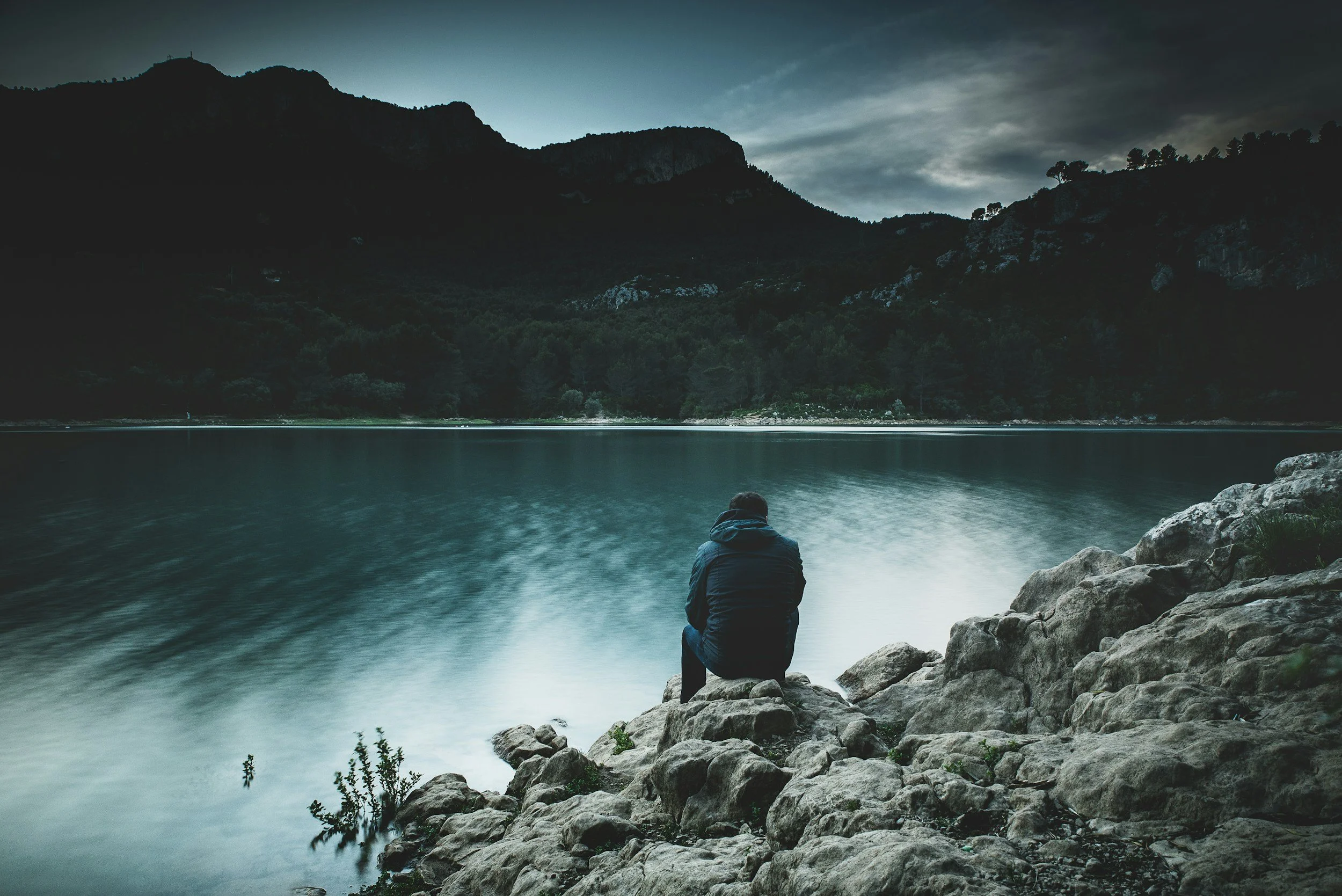 Person sitting on rocks by a lake with mountains and forest in the background during dusk or dawn.