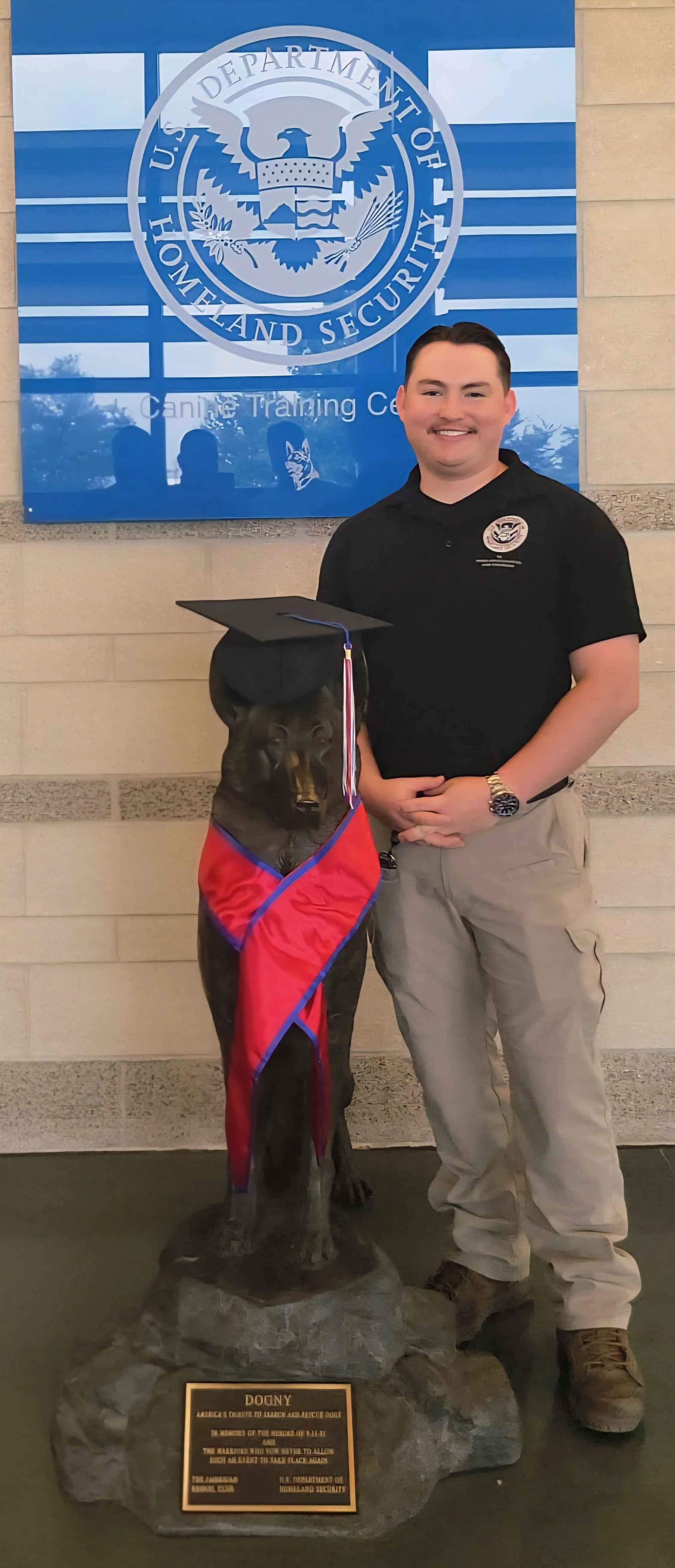 A man in a black uniform standing next to a black dog statue with a graduation cap and red cape. They are in front of a U.S. Department of Homeland Security sign, indicating a Canine Training Center. The man is smiling and wearing a watch and beige cargo pants.