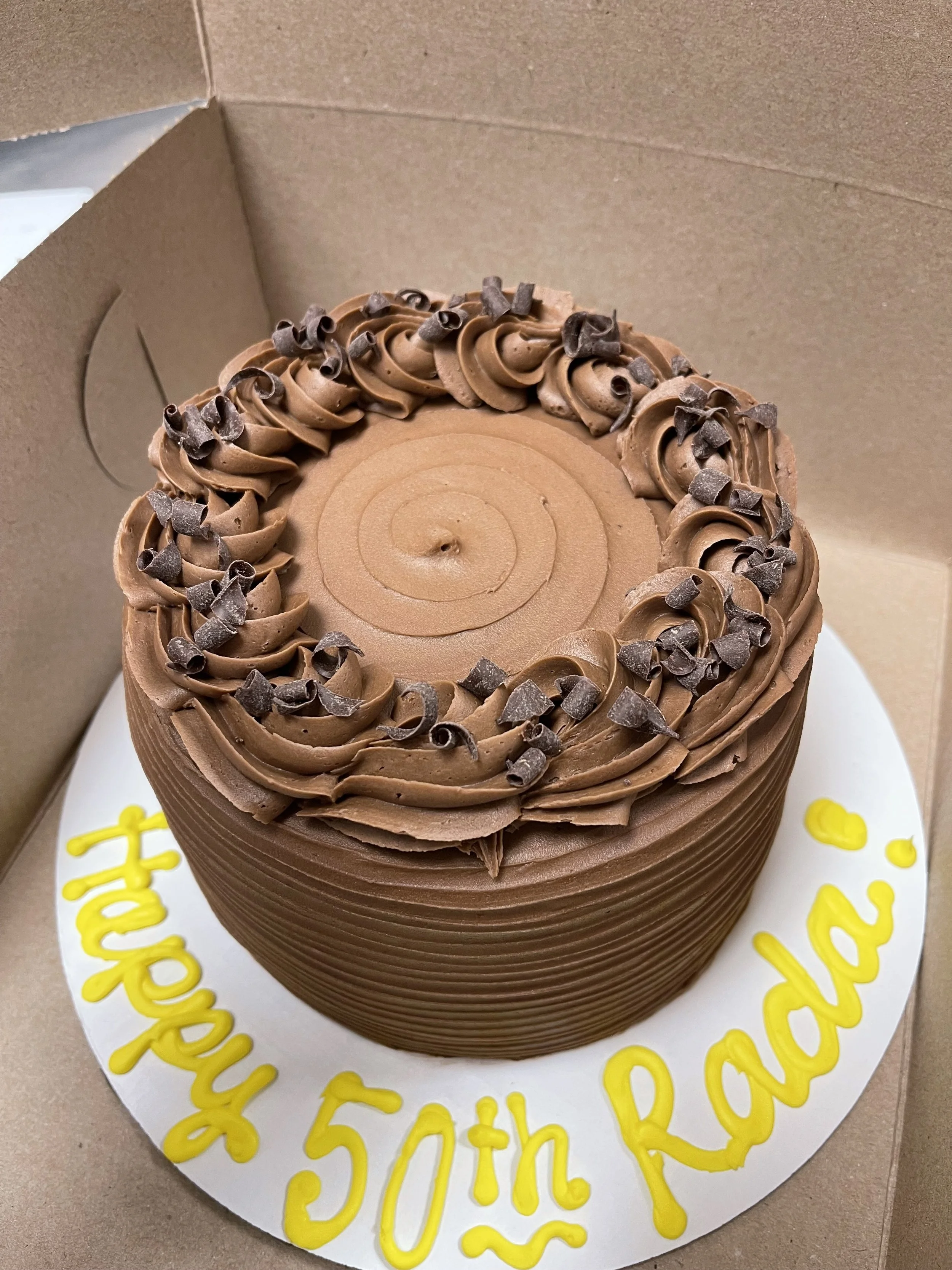 Chocolate birthday cake with spiral top decoration and chocolate curls, inside a cardboard box, with yellow writing on white cake board that says "Happy 50th Randa".