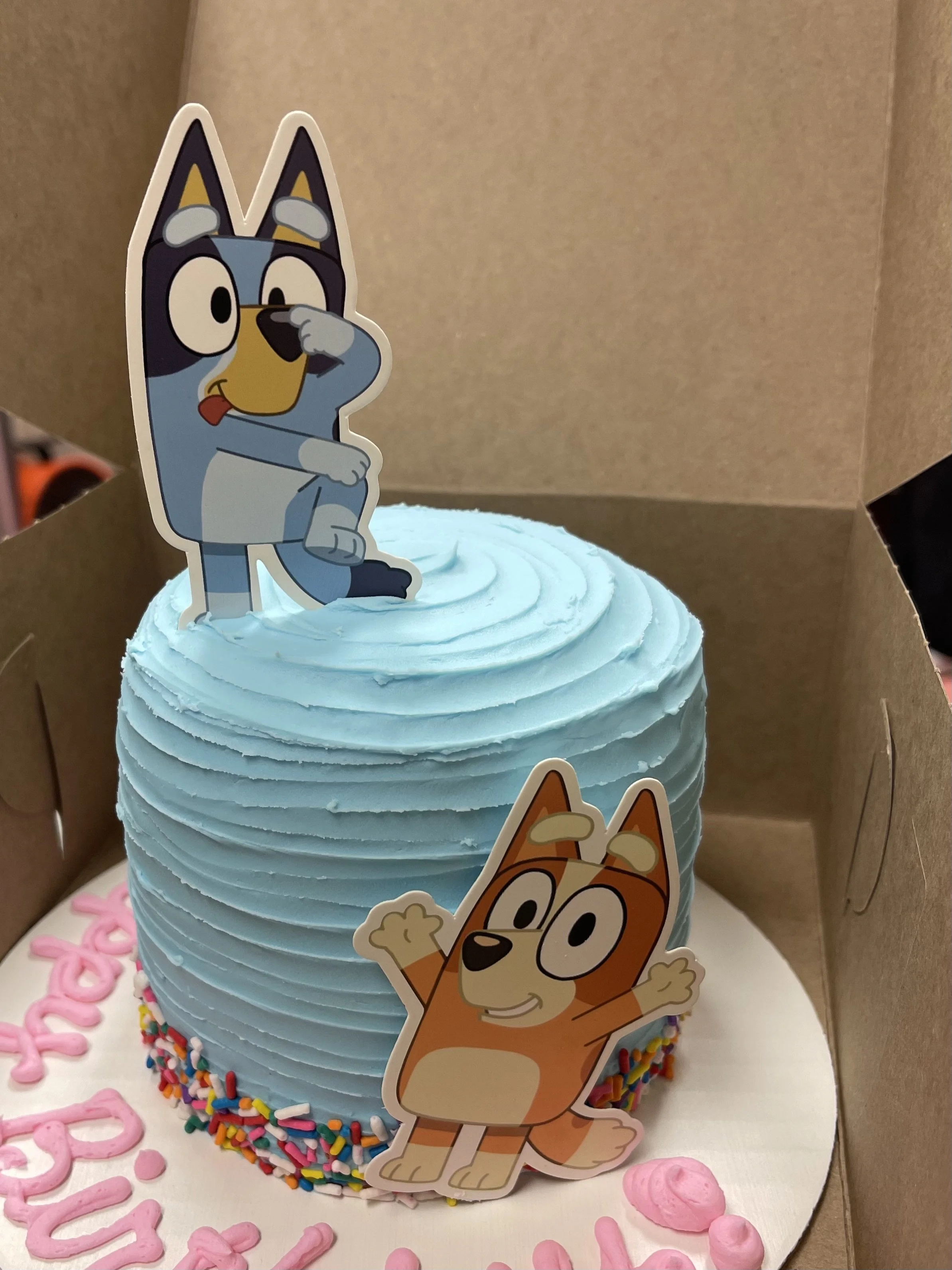A birthday cake with light blue frosting, decorated with colorful sprinkles, with cartoon character toppers of a blue dog with goggles and a brown dog with outstretched arms inside a cardboard box.