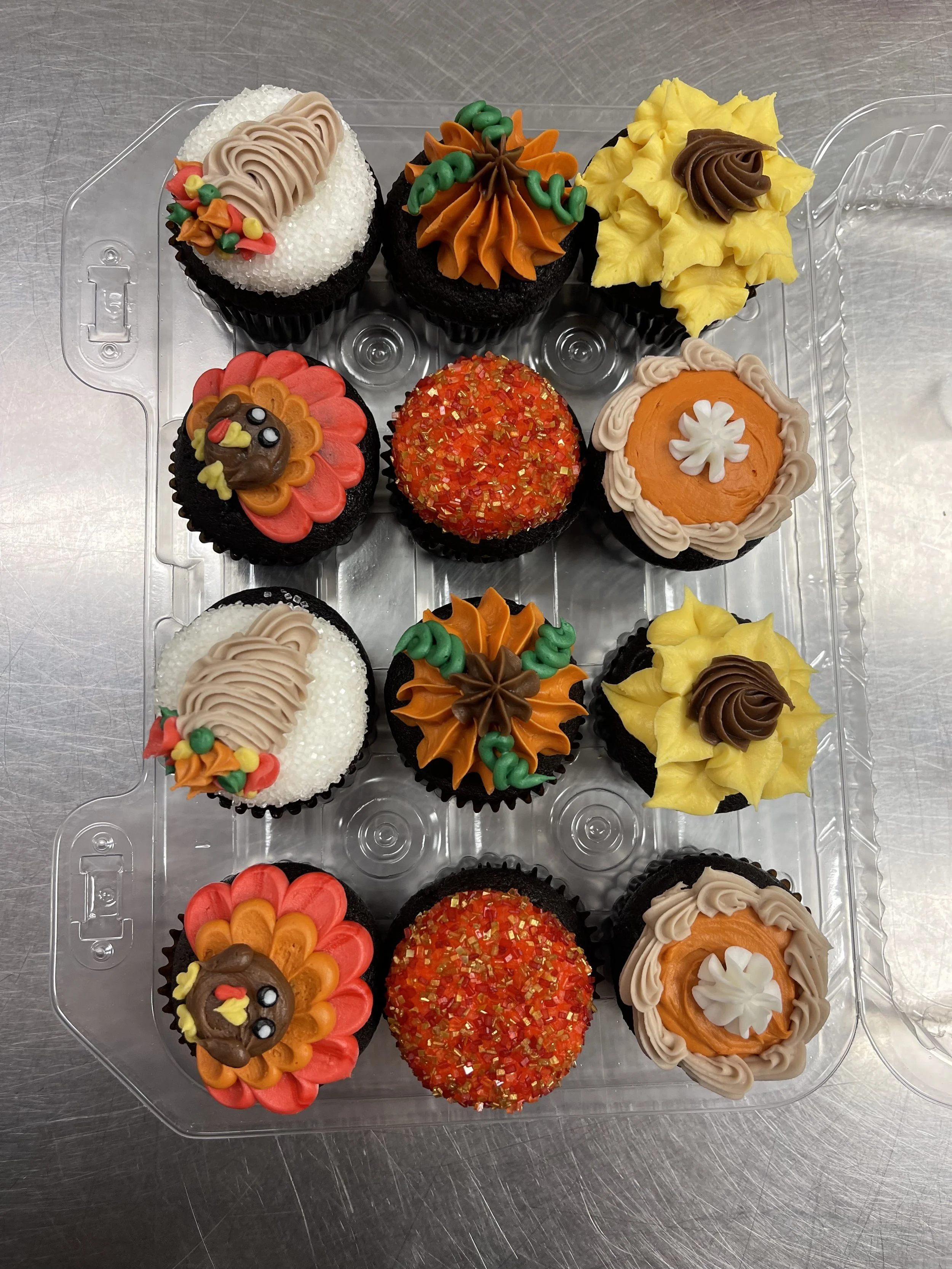 Assorted decorated cupcakes in a plastic tray with colorful frosting, sprinkles, and themed decorations.