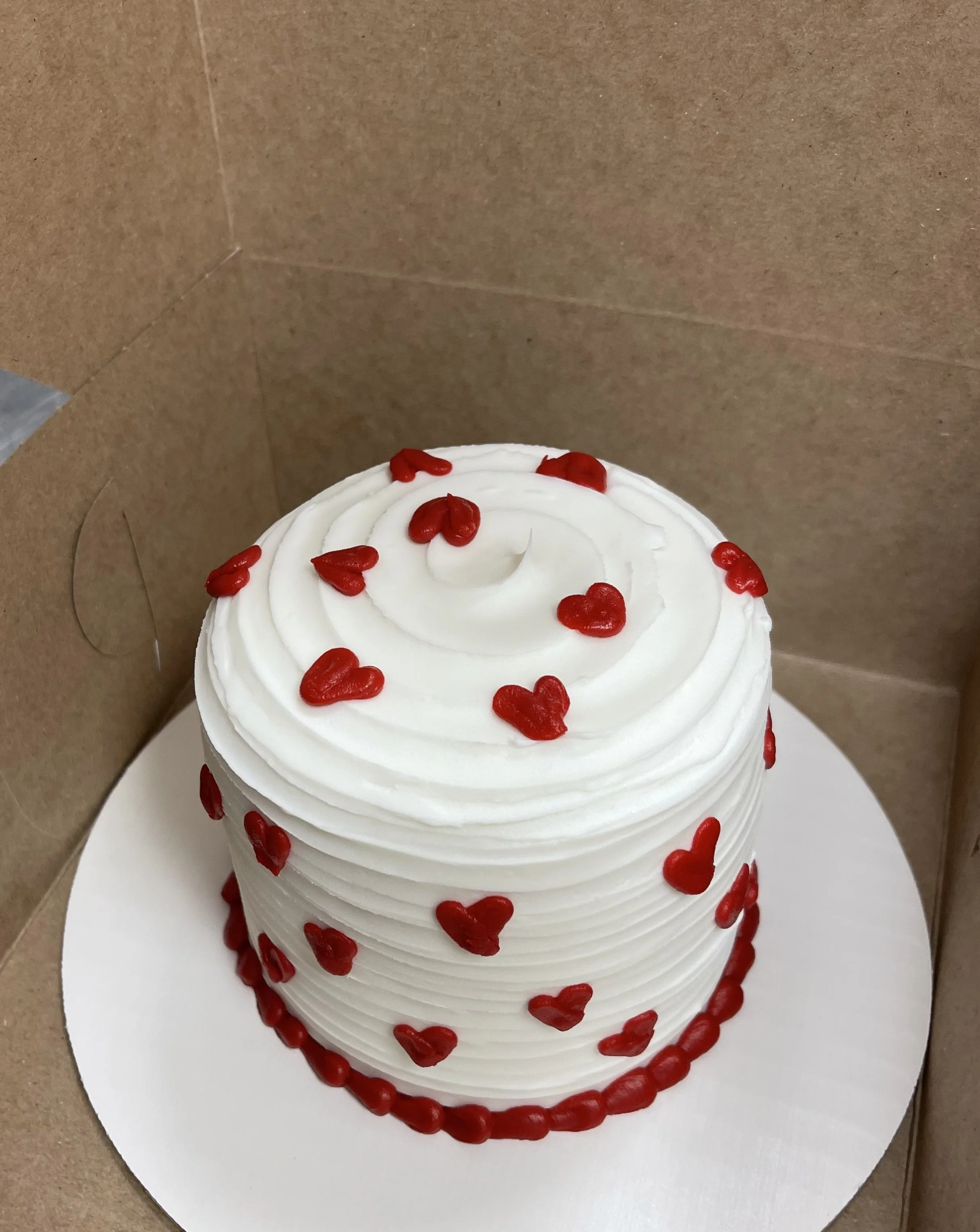 A white cake decorated with red heart-shaped sprinkles and a red border at the base, placed inside a cardboard box.