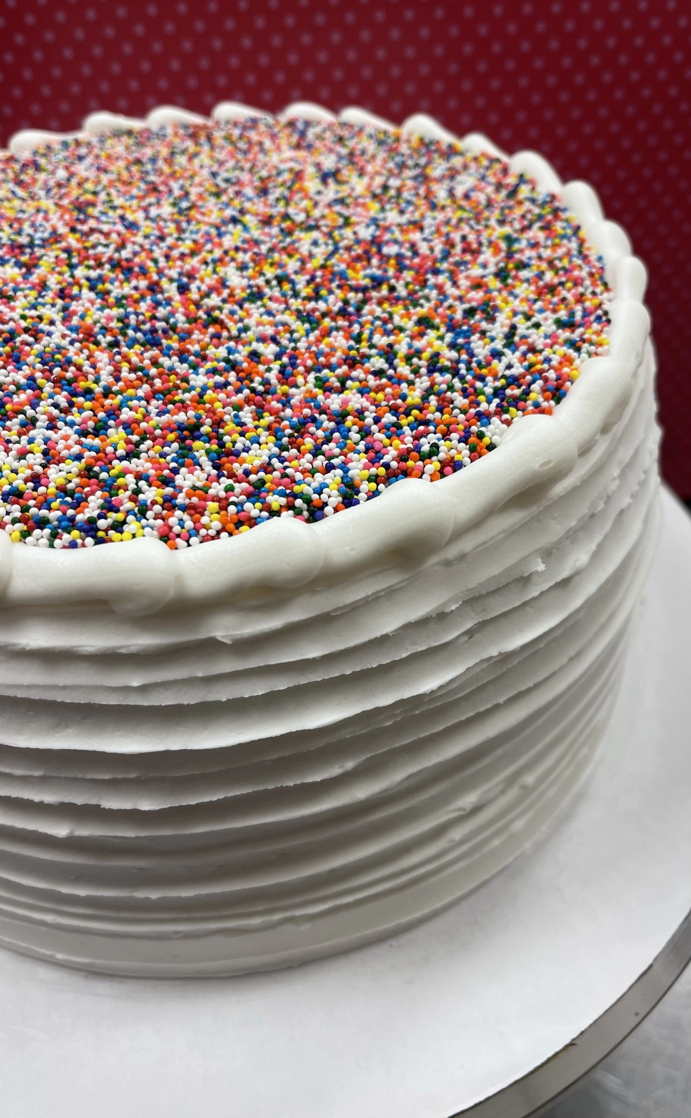 A round birthday cake with white frosting and colorful sprinkles on top, with decorative frosting edges, against a red polka dot background.