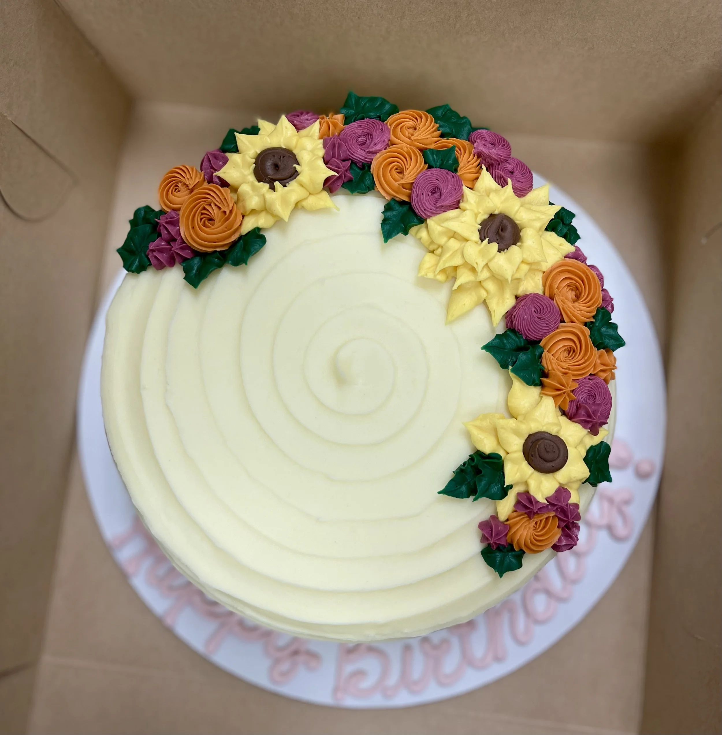A round cake with a smooth, creamy white surface decorated with colorful buttercream flowers including yellow sunflowers with brown centers, orange roses, and purple roses, with green leafy buttercream accents. The cake is in a cardboard box, and a p