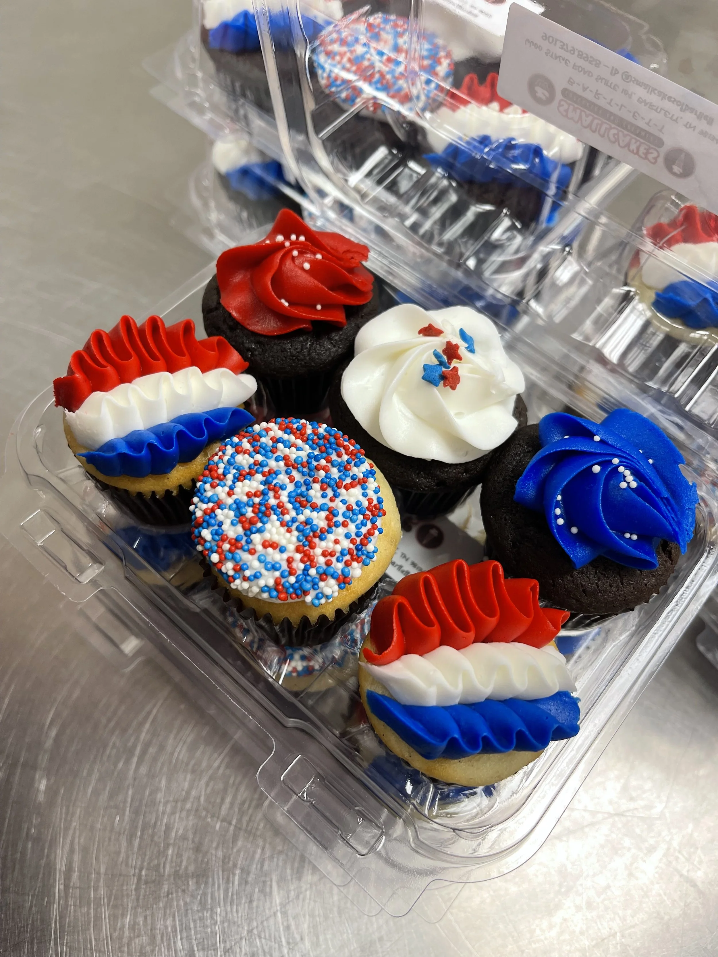 A clear plastic container holding decorated cupcakes with red, white, and blue themed frosting and sprinkles, ready for patriotic celebration.