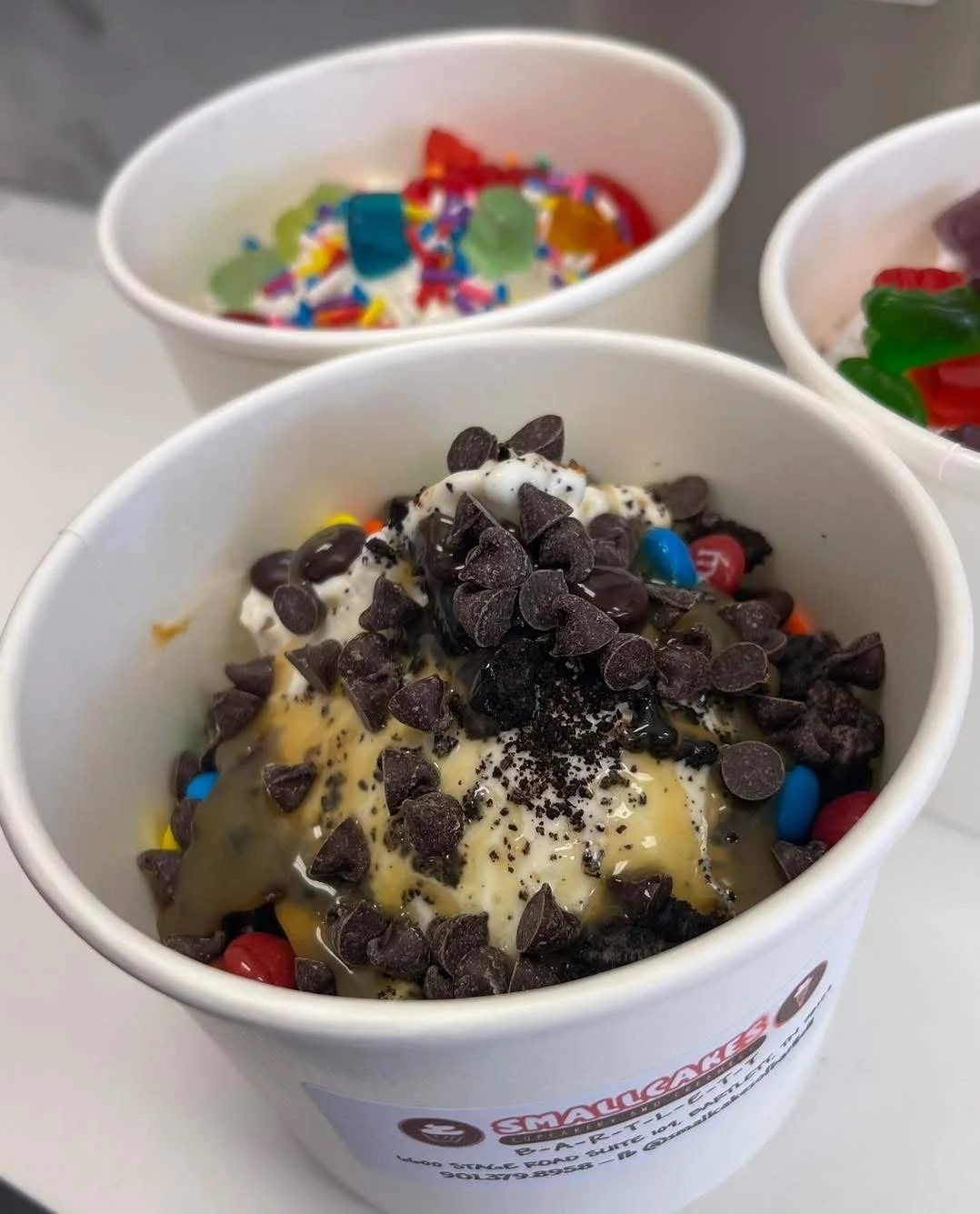 Close-up of a cup filled with vanilla ice cream topped with chocolate chips, colorful candy-coated chocolates, and chocolate syrup, with two additional cups of ice cream with sprinkles and gummy candies in the background.