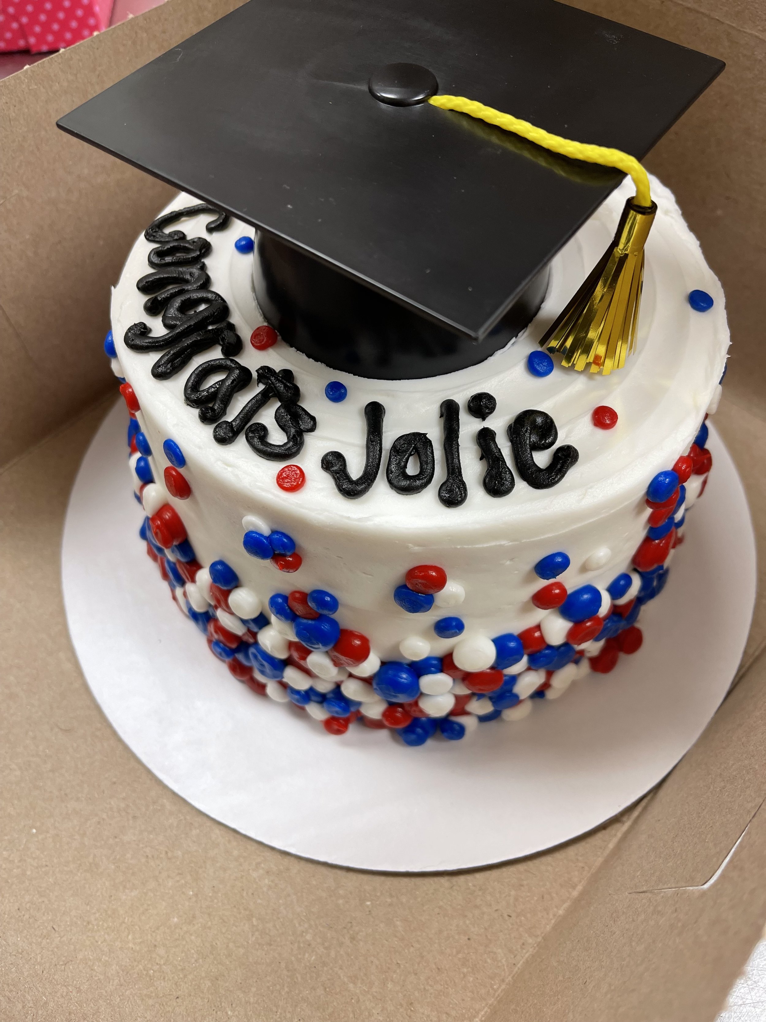 A graduation cake decorated with white, red, and blue sprinkles, with a black graduation cap and a gold tassel on top. The words 'Congrats Jorie' are written in black icing on the cake.