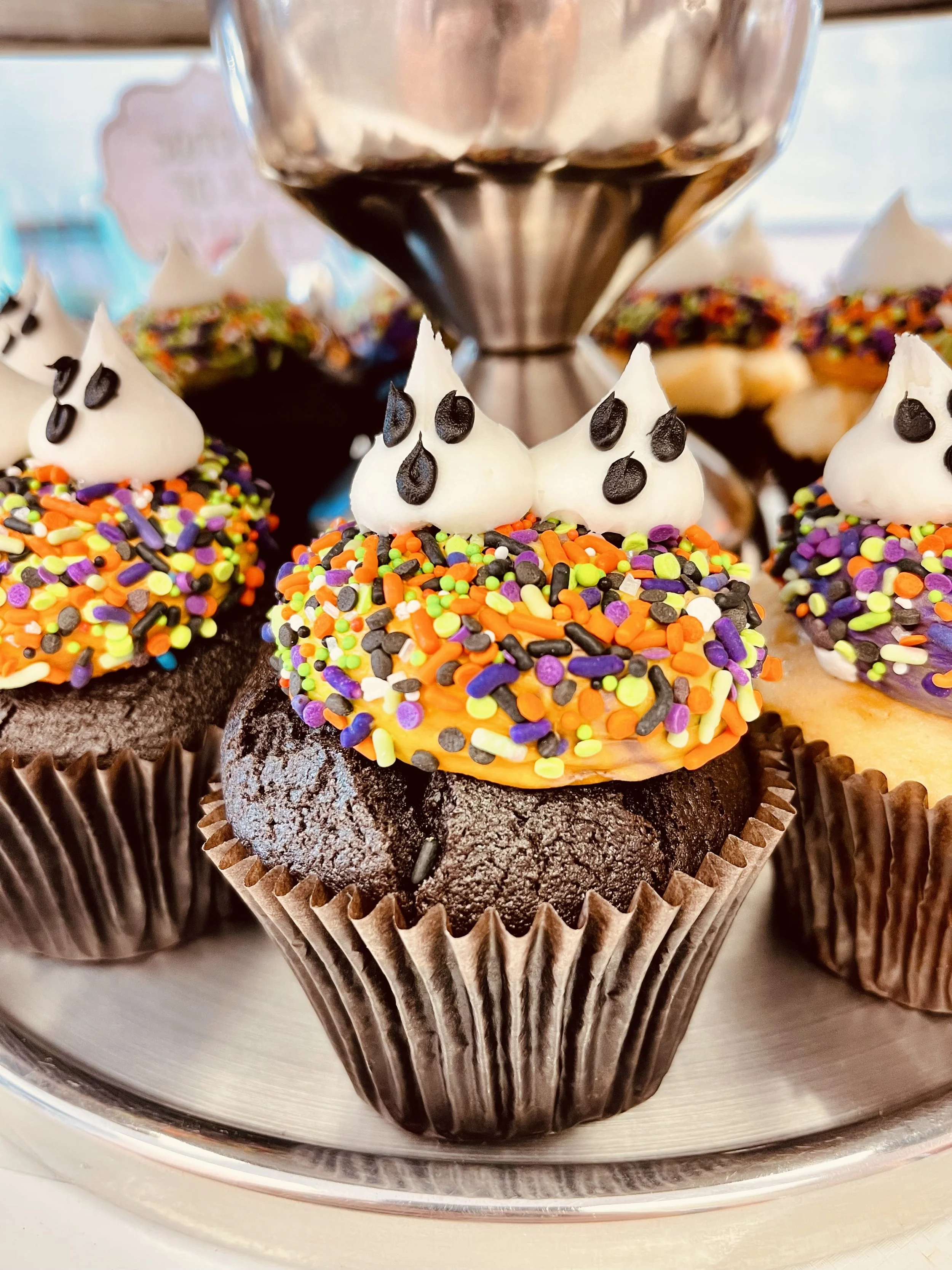 Cupcake with black cake base, topped with white frosting dots resembling ghost faces, and colorful sprinkles.