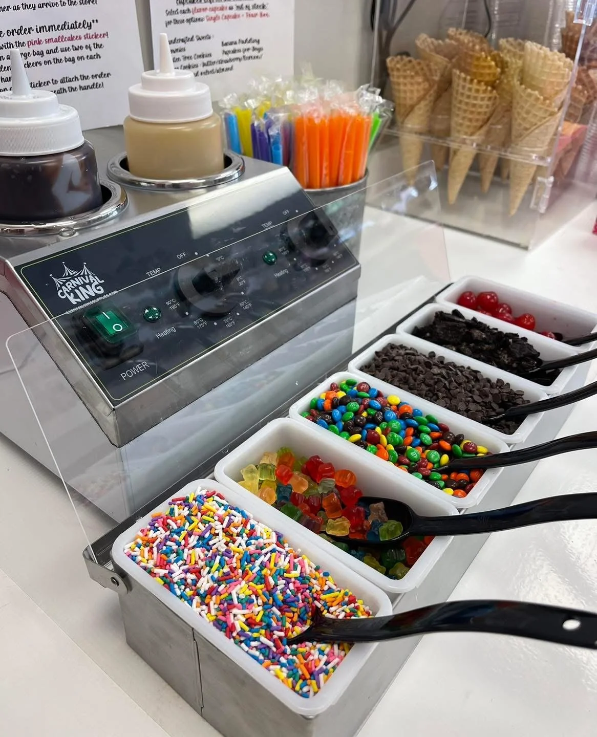 A topping station with various toppings for ice cream, including rainbow sprinkles, gummy bears, chocolate chips, chocolate crumbs, and maraschino cherries, along with syrup dispensers and waffle cones in a plastic holder.