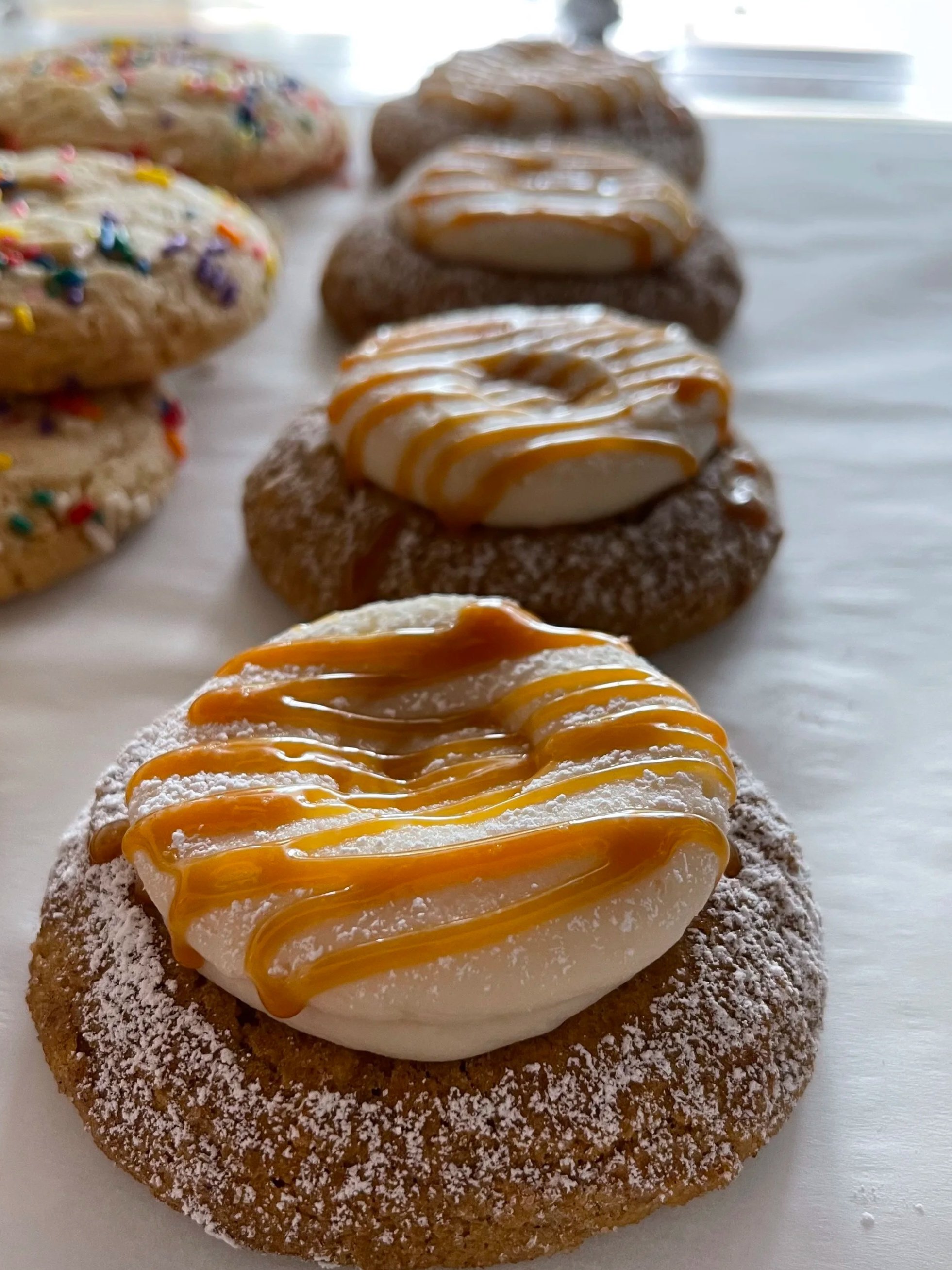 Close-up of a row of decorated cookies, with the front cookie topped with white frosting, caramel drizzle, and powdered sugar.
