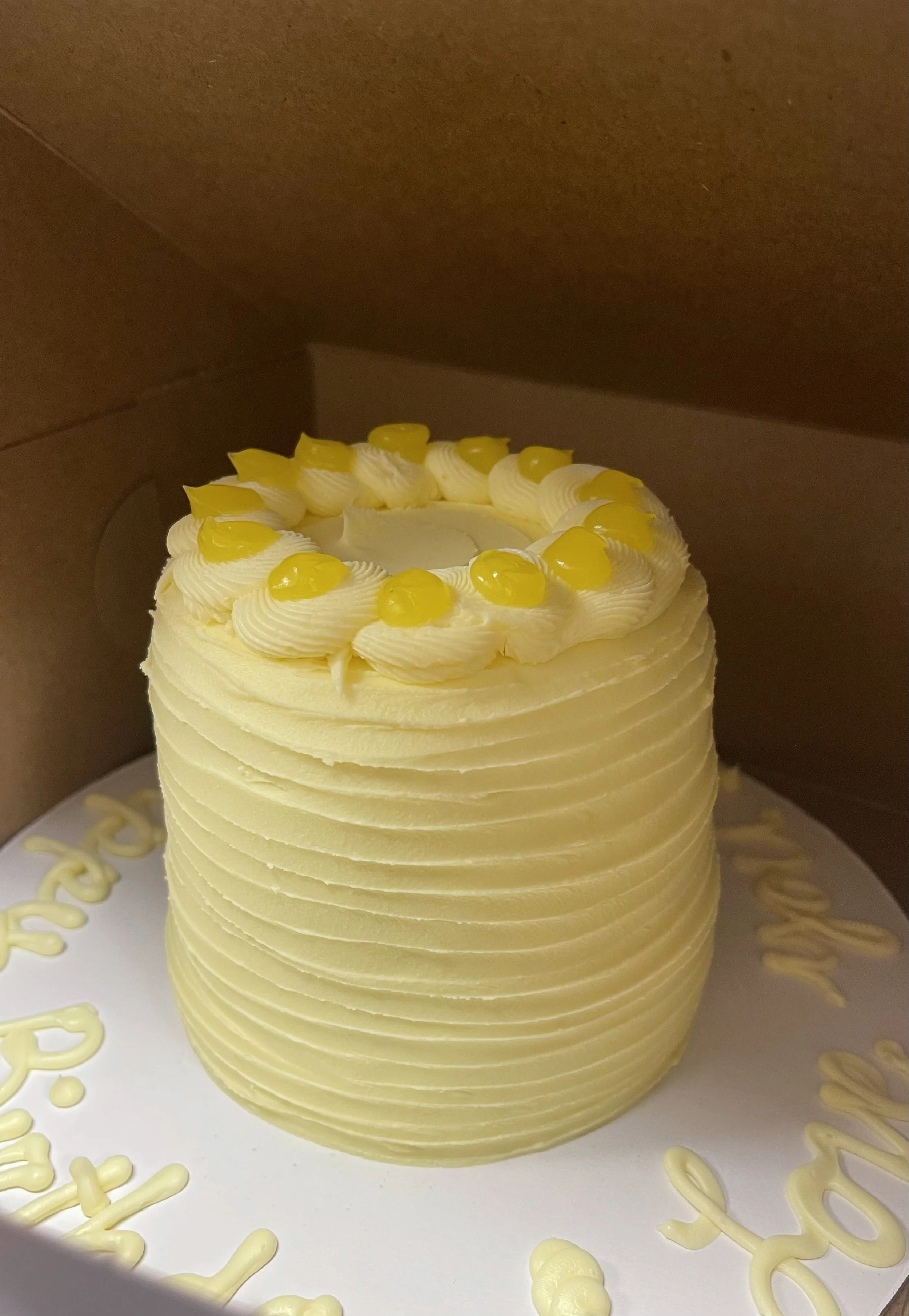 A tall white birthday cake decorated with yellow and white icing in a swirl pattern, topped with yellow icing dots, inside a brown cardboard box.