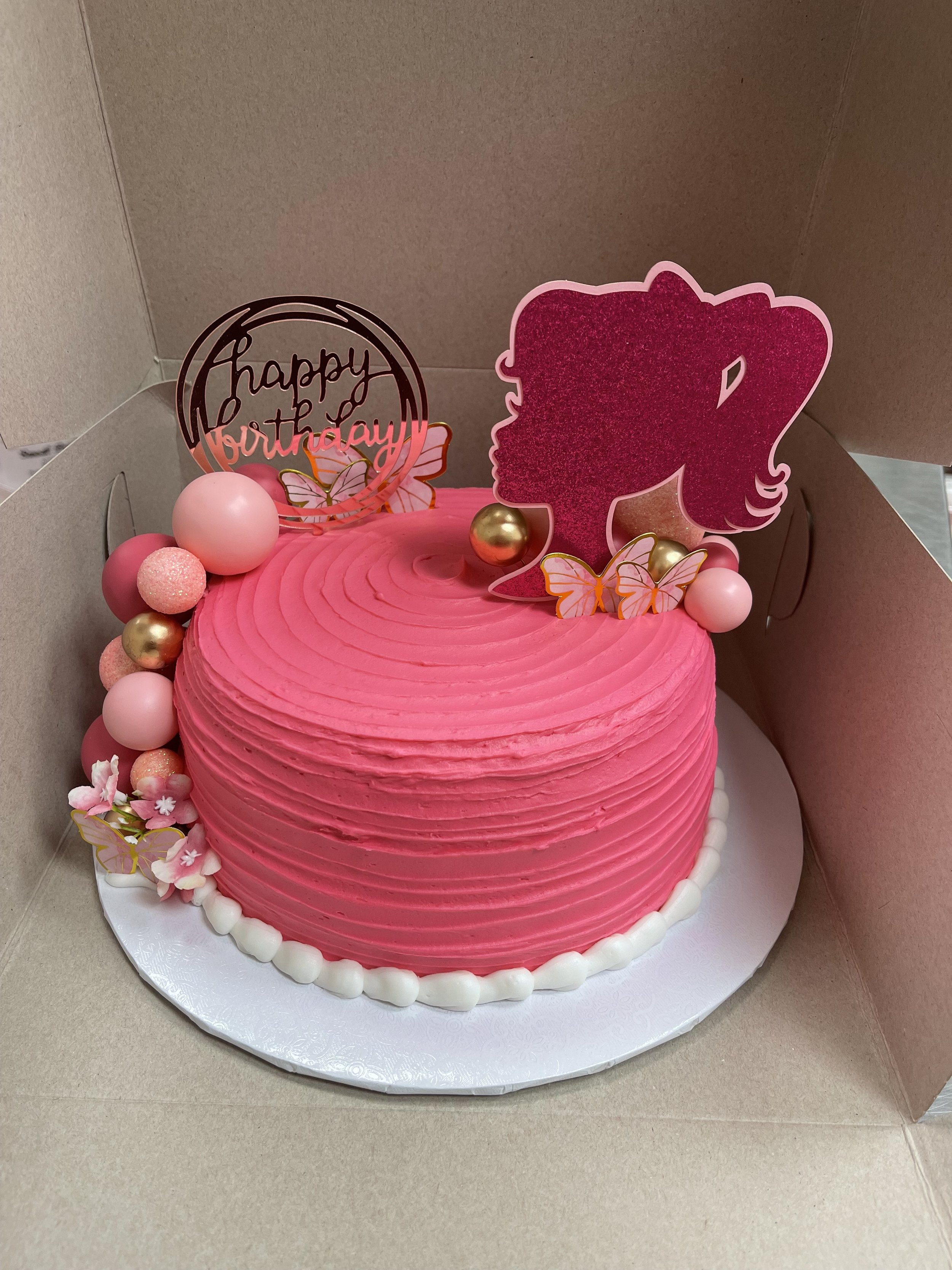 Pink, heart-shaped birthday cake with decorations, flowers, butterflies, and 'happy birthday' topper inside a cardboard box.