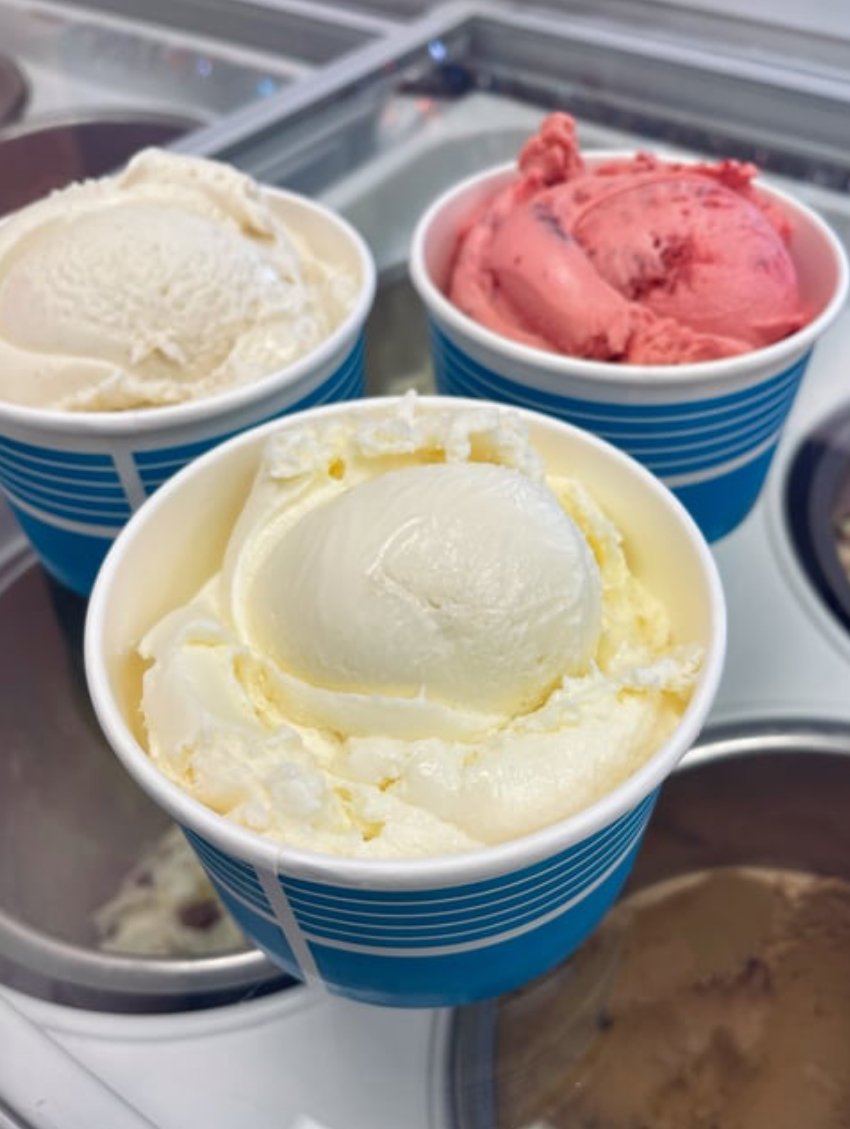 Three cups of ice cream in different flavors: vanilla, strawberry, and lemon, on a glass display freezer.