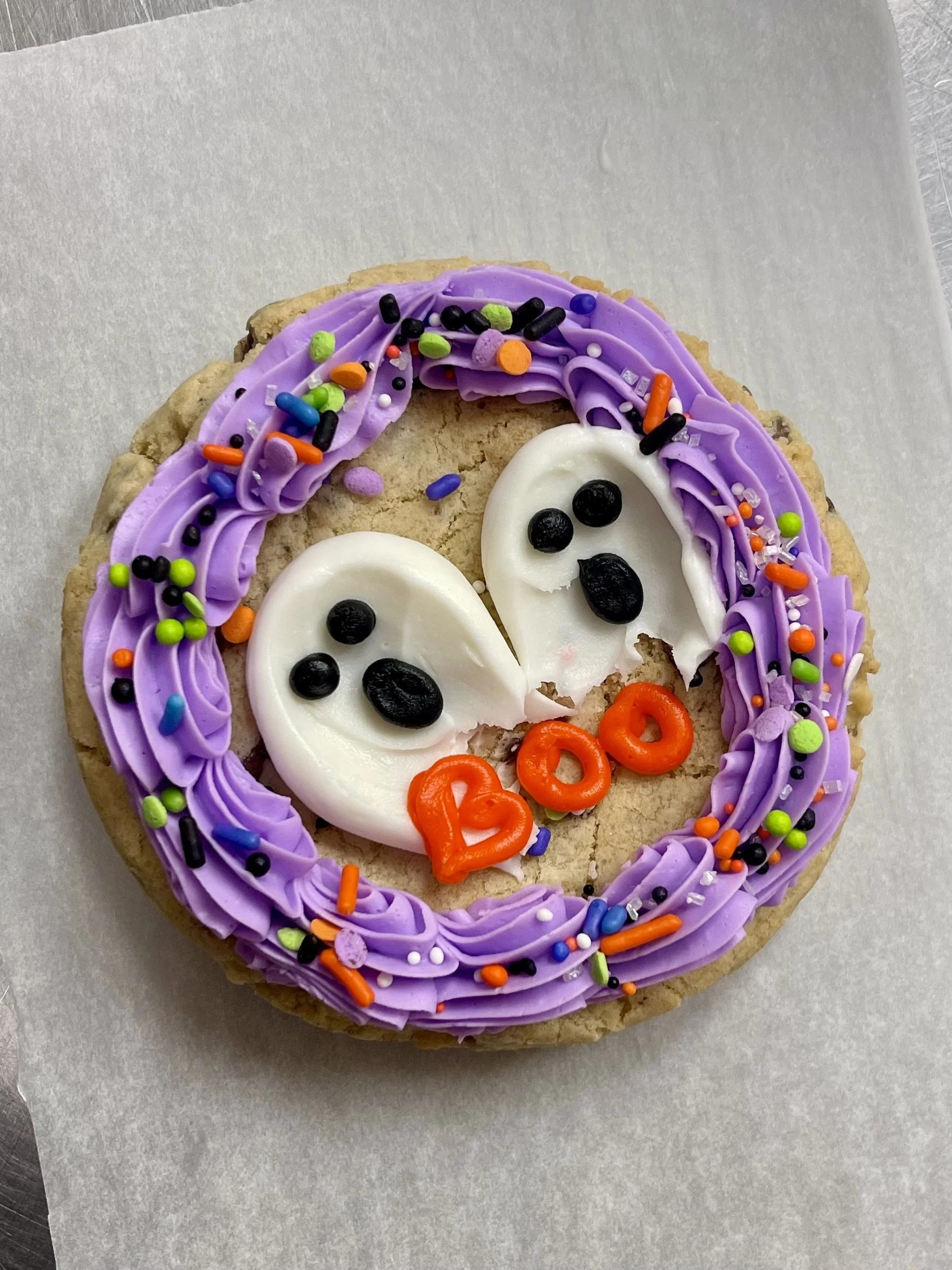 Decorative Halloween cookie with purple icing, colorful sprinkles, two ghost faces made of white icing with black eyes and mouths, and orange "BOO" lettering.