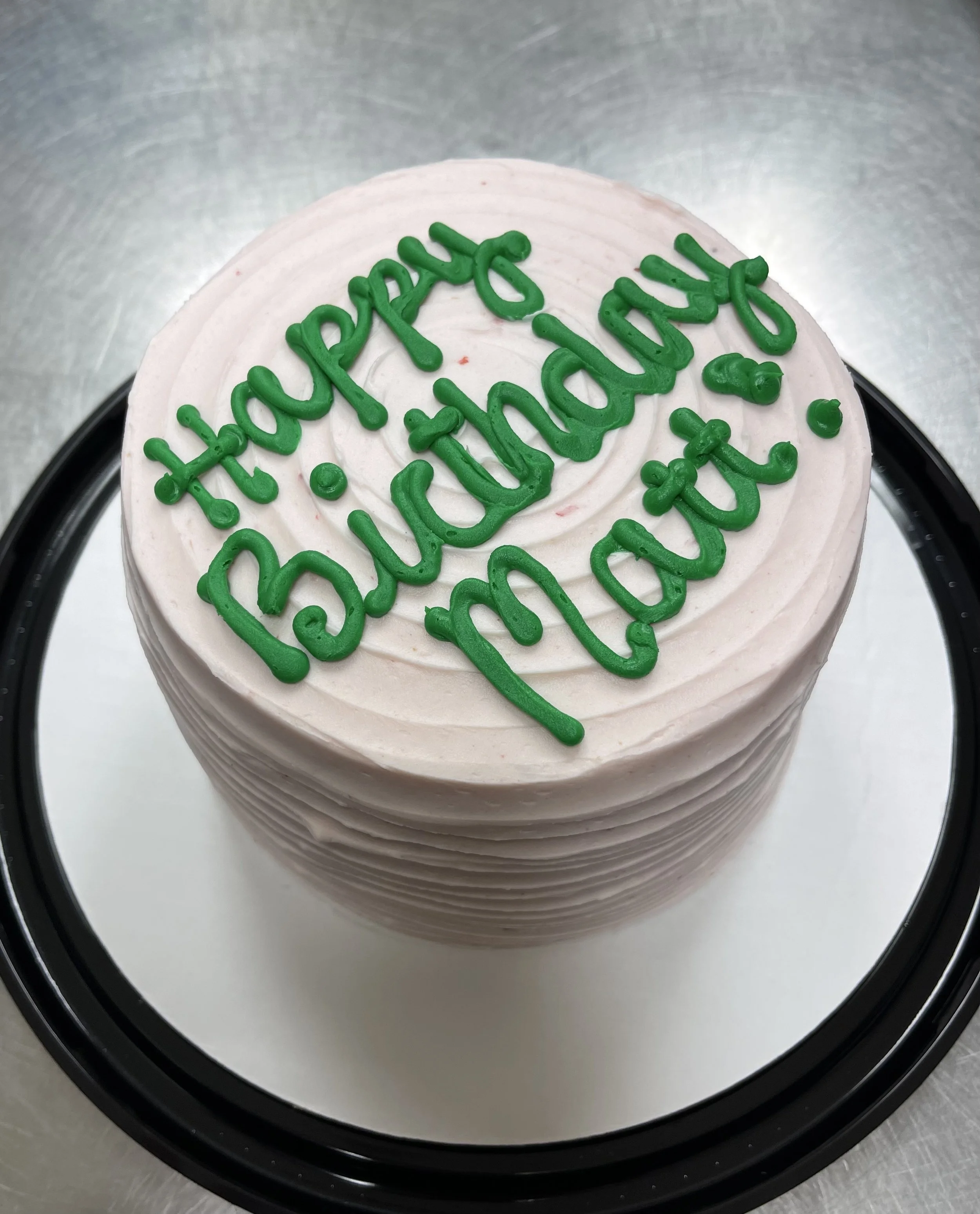 Pink birthday cake with green icing message that says 'Happy Birthday, Maddie' on top, placed on a black circular cake stand.