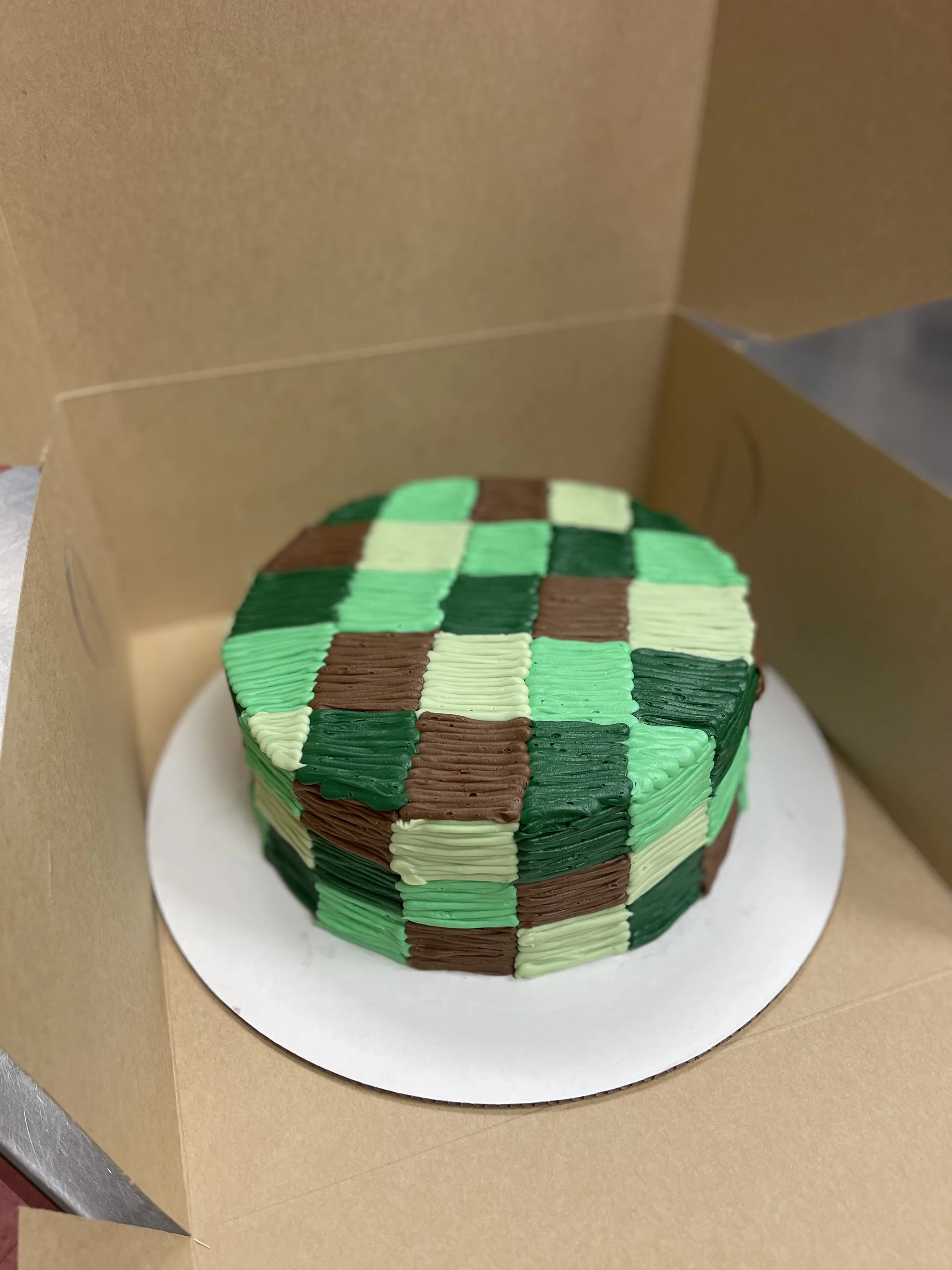 Decorative cake with a pixelated camouflage pattern in green, brown, and tan colors inside a cardboard box.
