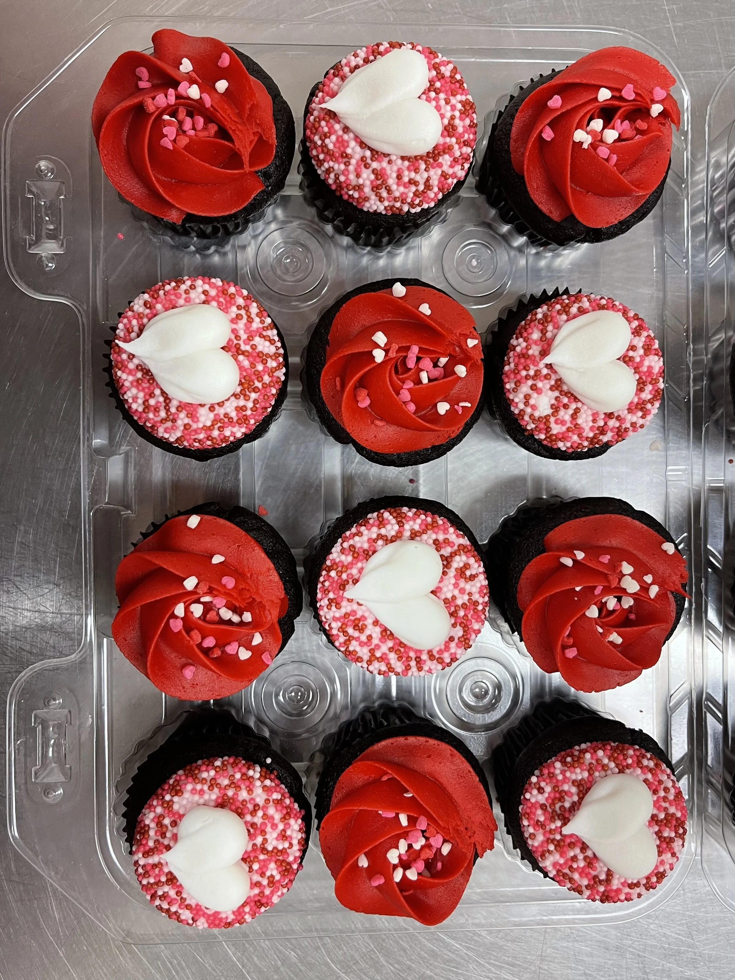 A tray of cupcakes decorated with red and white icing, heart-shaped sprinkles, and white heart-shaped toppers for Valentine's Day.
