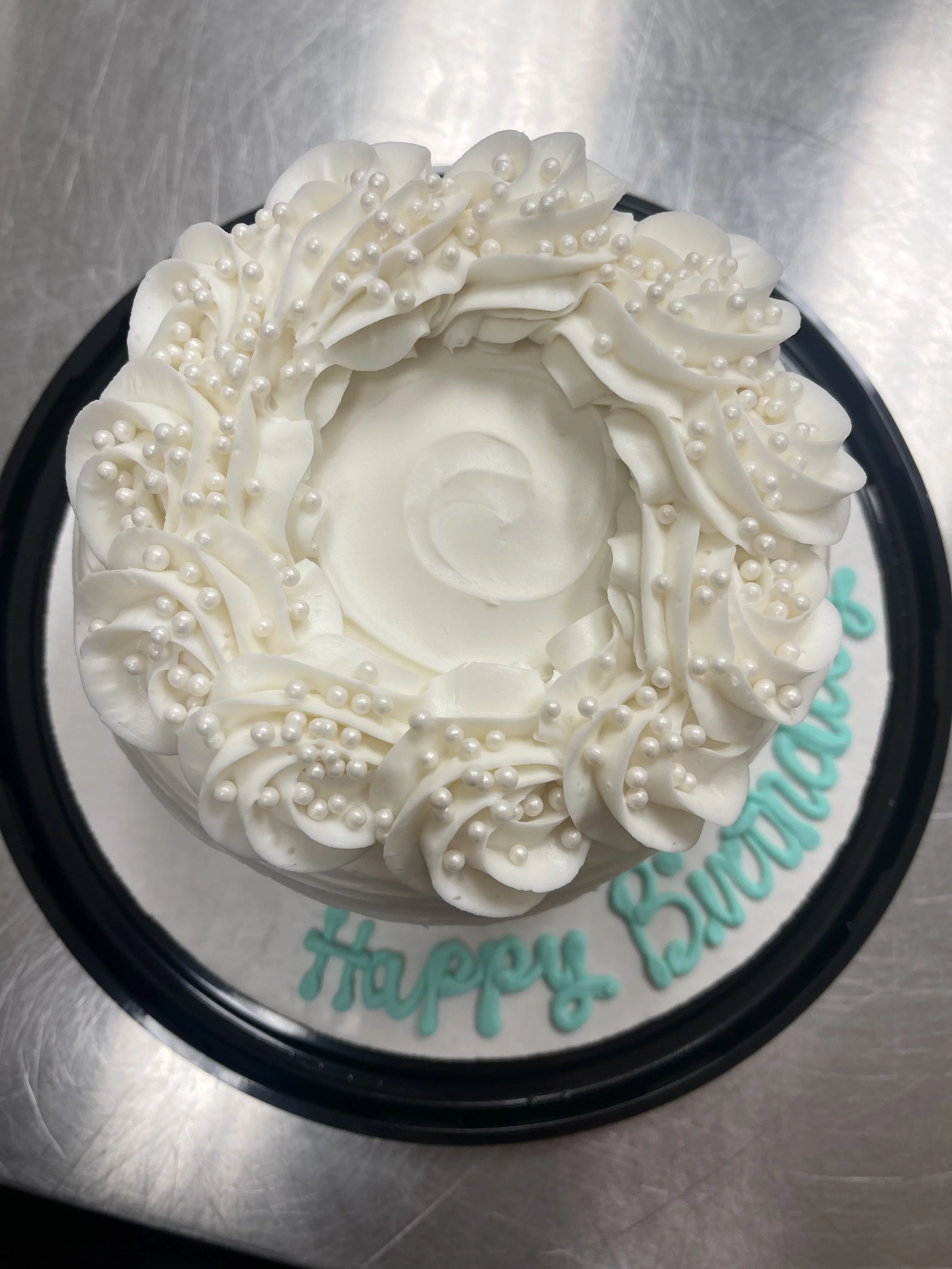 White birthday cake with piped frosting and pearl decorations, on a black tray with a "Happy Birthday" message written in blue on the surface.