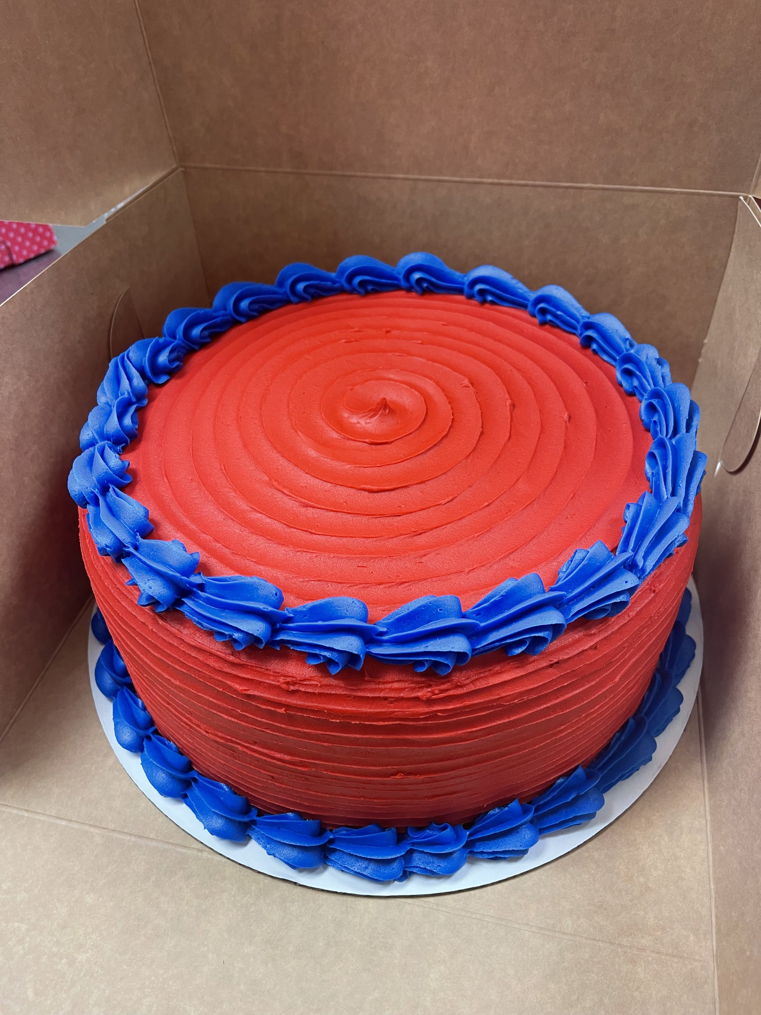 Red birthday cake with blue frosting accents and a swirl pattern on top, inside a cardboard box.