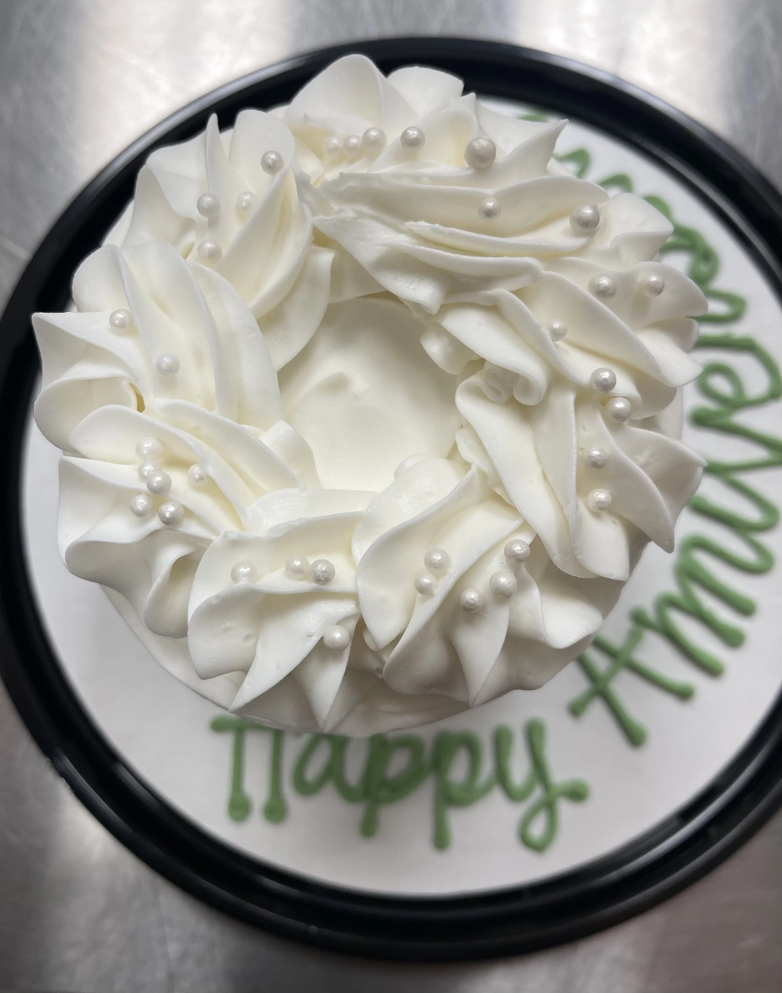 A round cake with white frosting decorated with piped rosettes and pearl-like sprinkles, with a black border around the top edge and the words 'Happy Anniversary' written in green around the base.
