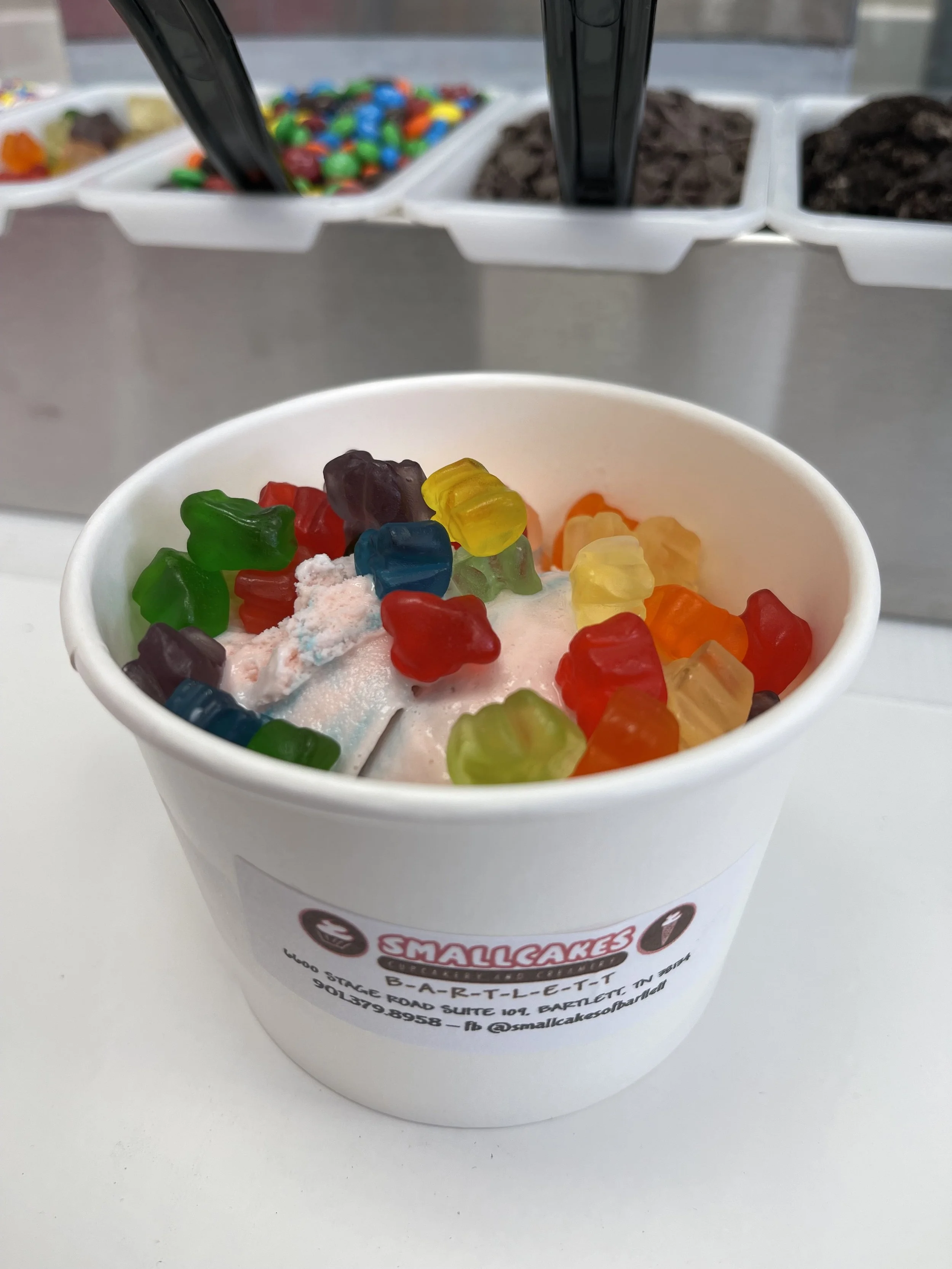 Cup of ice cream topped with colorful gummy bears, with toppings station including candies and cookies in the background.