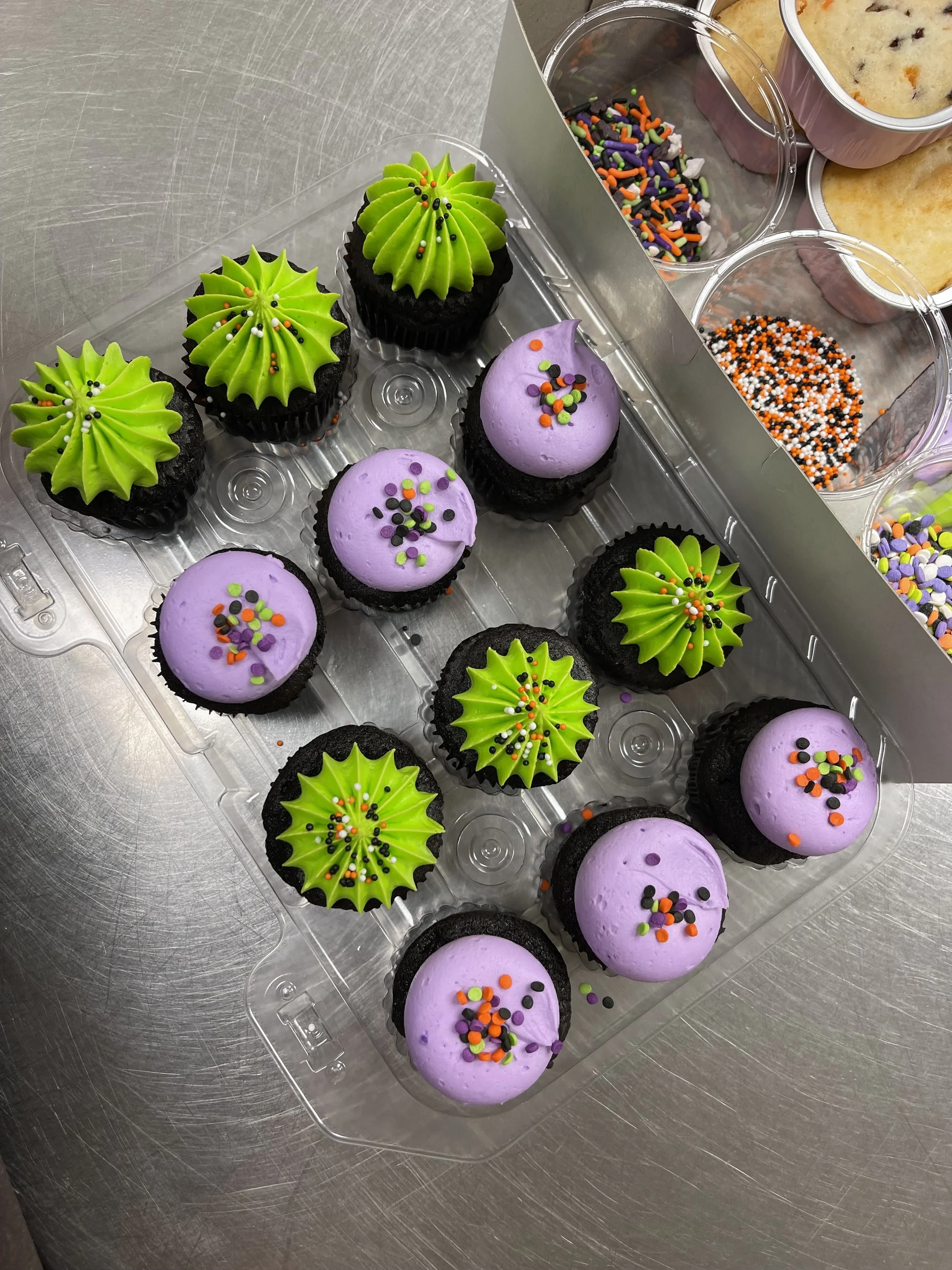 Cupcakes decorated with green and purple frosting, topped with rainbow sprinkles, arranged in a plastic container with Halloween-themed decorations.