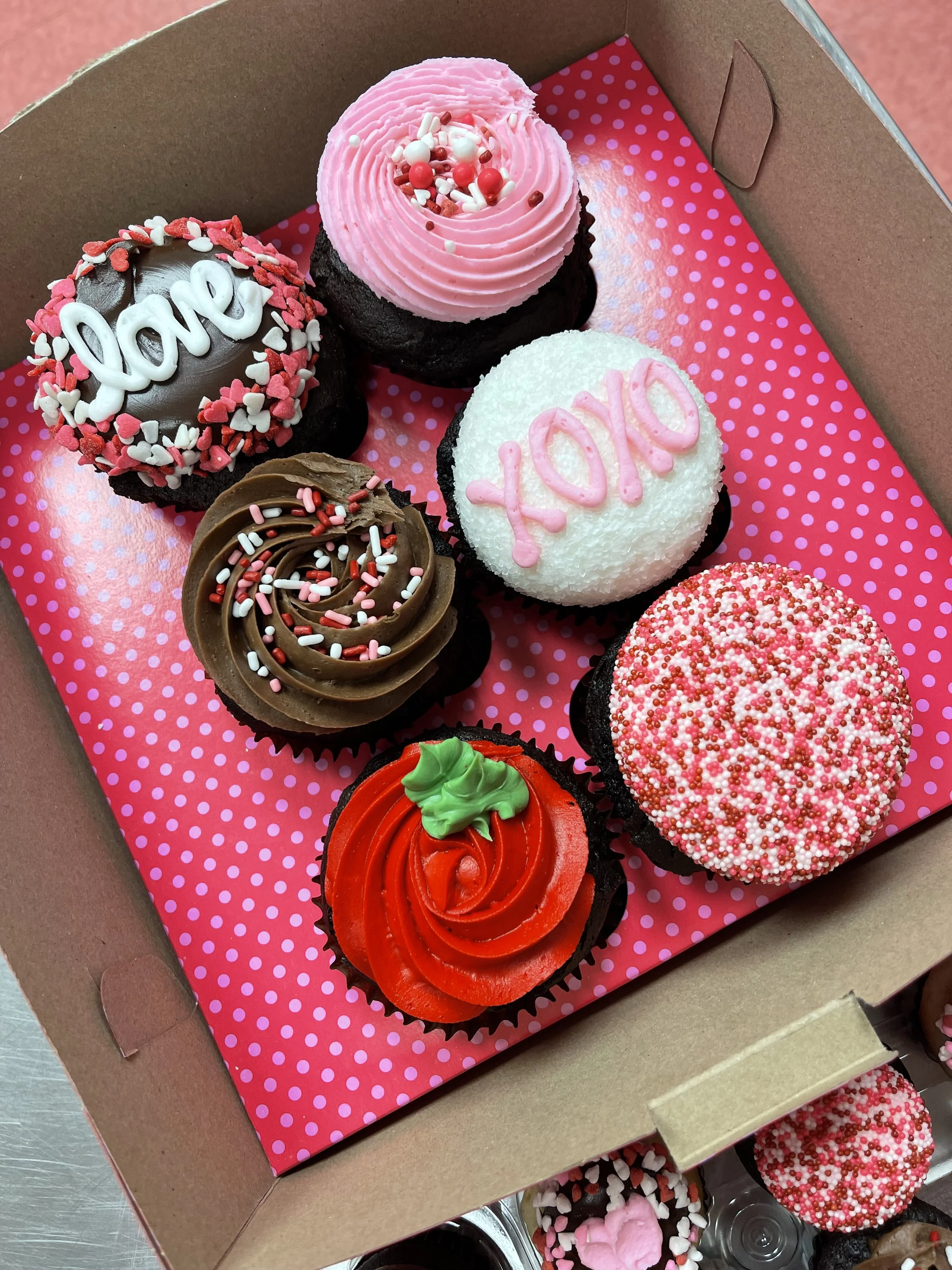 Box of Valentine's Day cupcakes with pink, red, and white frosting and sprinkles, decorated with love and XO words.