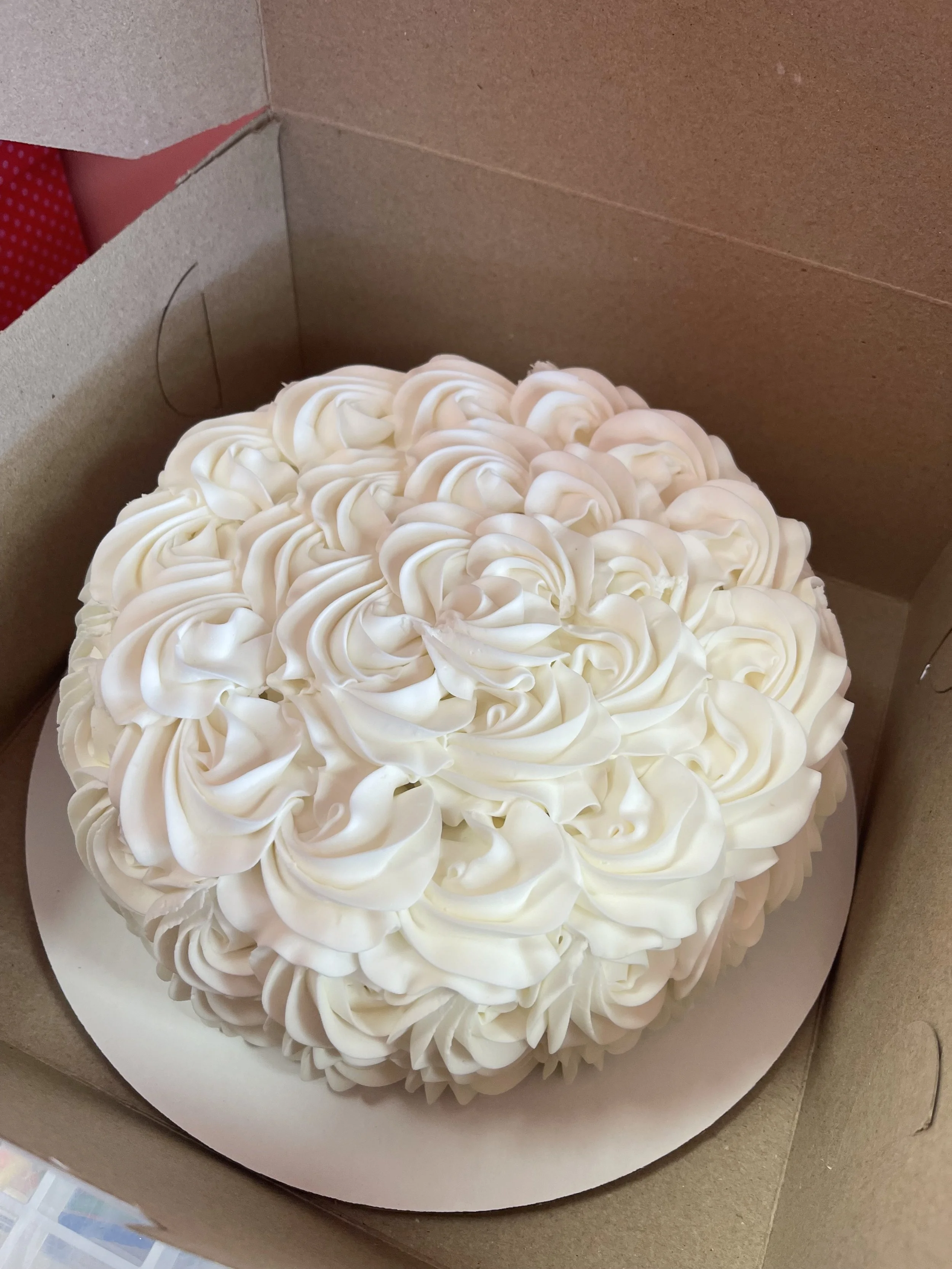 A round cake decorated with white buttercream frosting in a ruffled pattern inside a cardboard box.