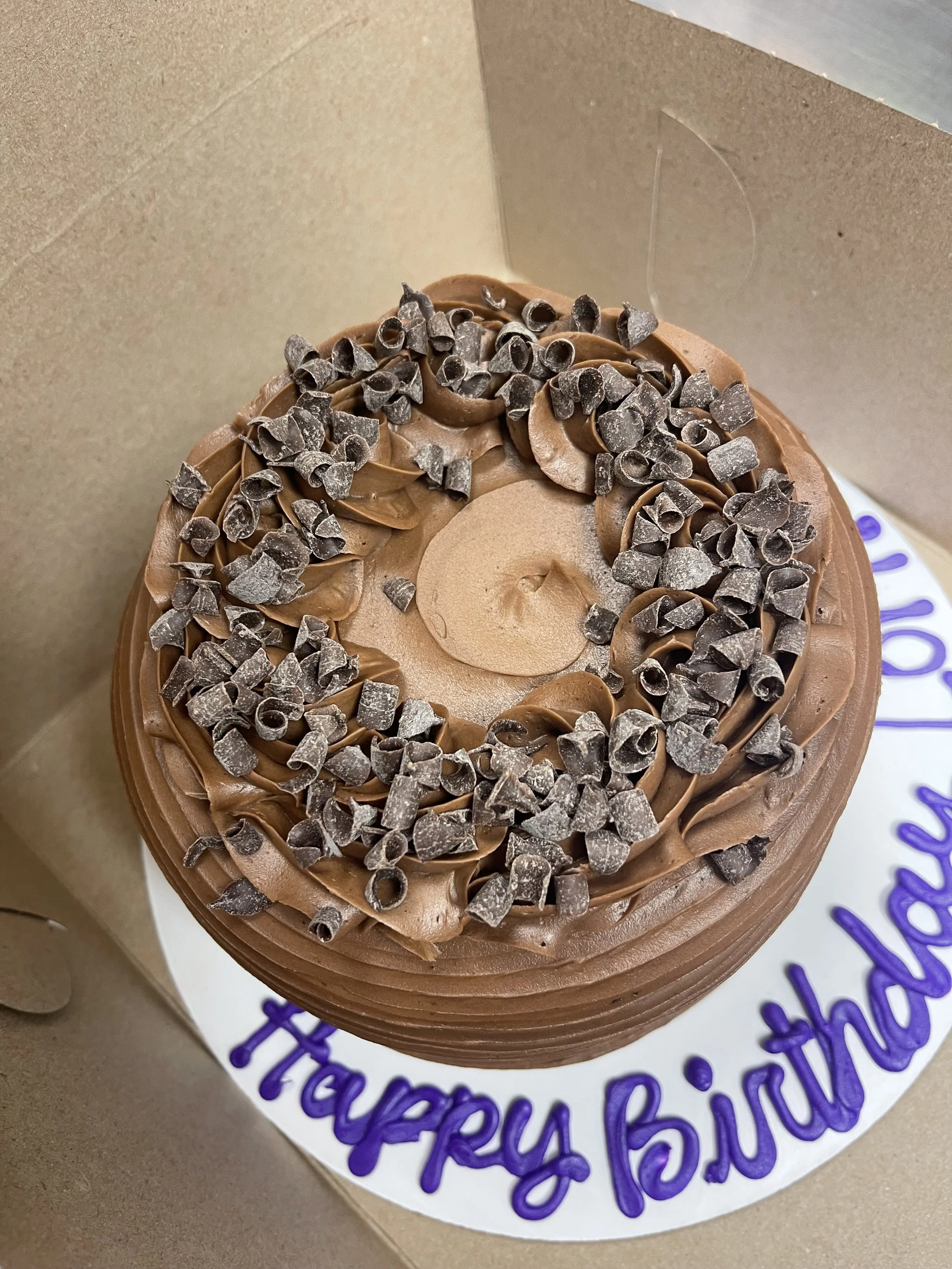 Round chocolate birthday cake with chocolate shavings on top, placed on a white cake board with 'Happy Birthday' written in purple. The cake is in a cardboard box.