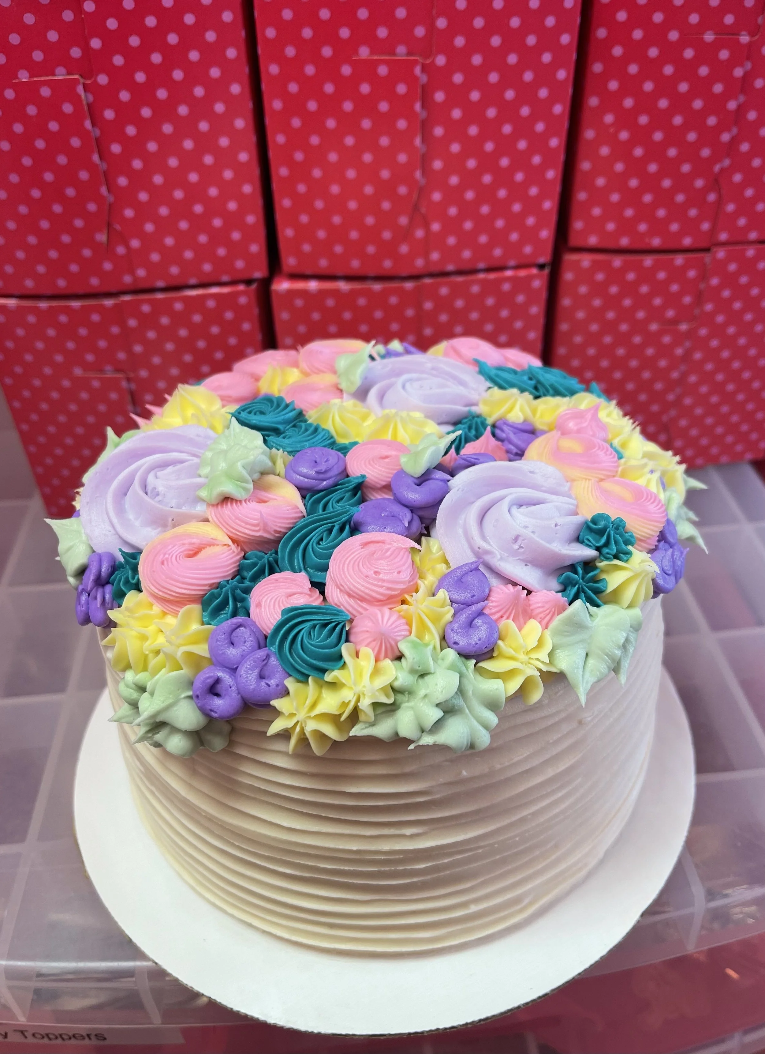 Decorated birthday cake with purple, pink, blue, yellow, and green frosting flowers on top, in front of pink and white polka dot boxes.