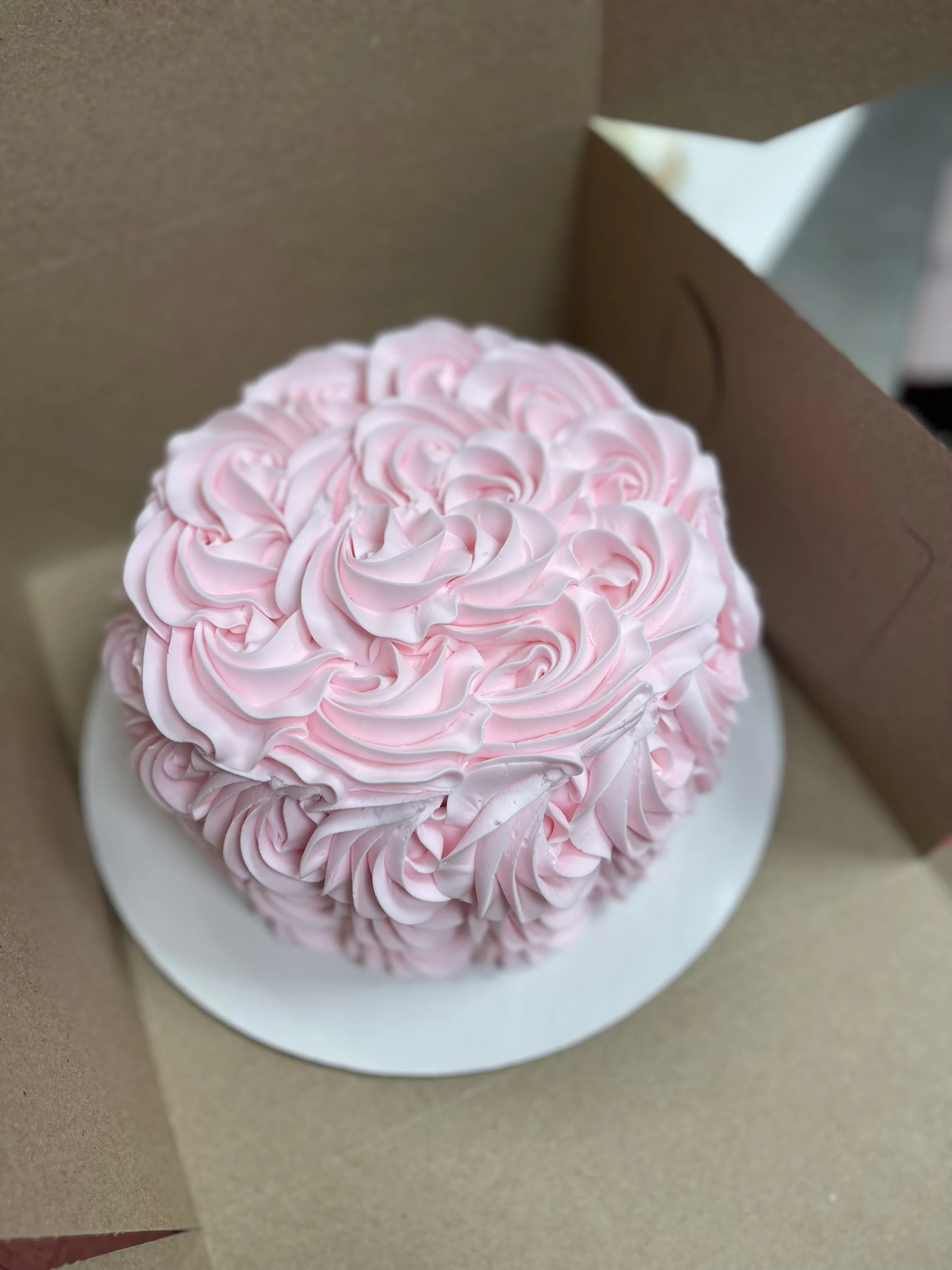 Round cake with pink frosting piped in rosette pattern inside a cardboard box on a white cake board.