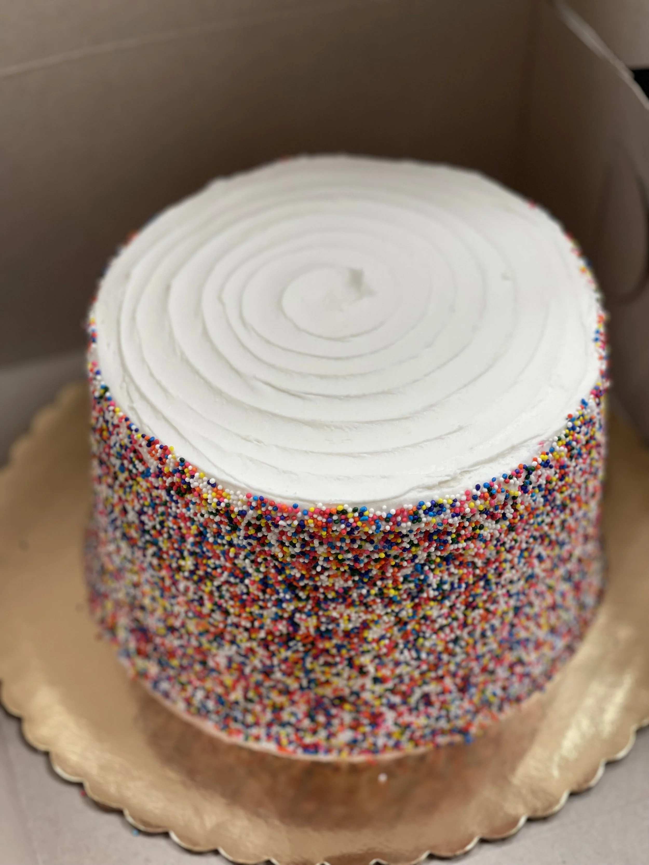 Round cake with white frosting, decorated with multicolored sprinkles on the sides, in a cardboard box on a gold-colored base.