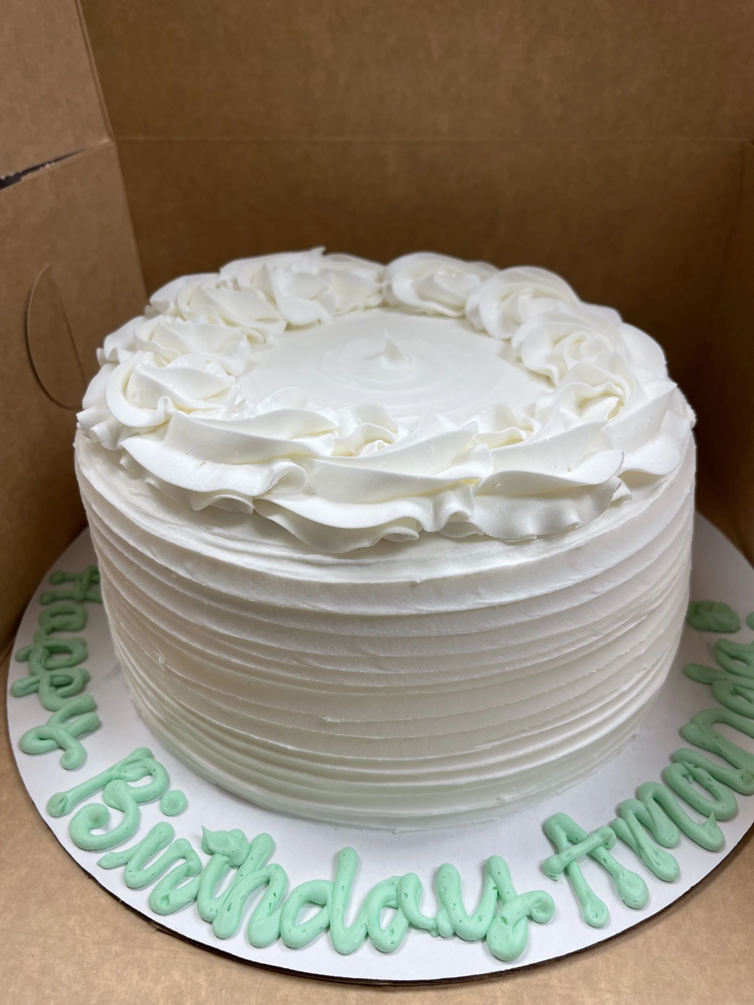 White birthday cake with decorative frosting on top in a cardboard box, with green and white words on the cake board reading 'Happy 38th birthday Touche'.