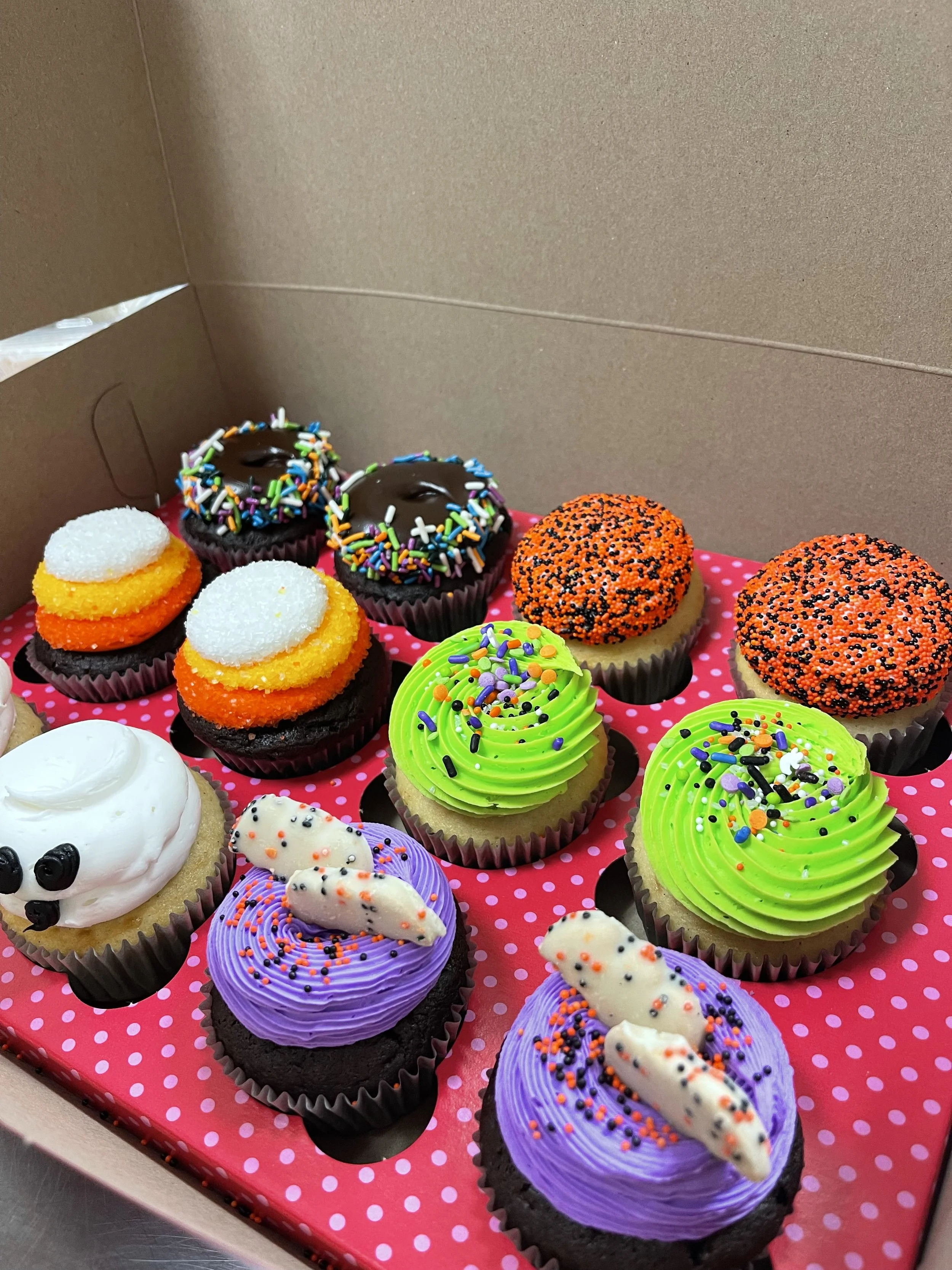 A box of colorful Halloween-themed cupcakes decorated with sprinkles, frosting, and various toppings.