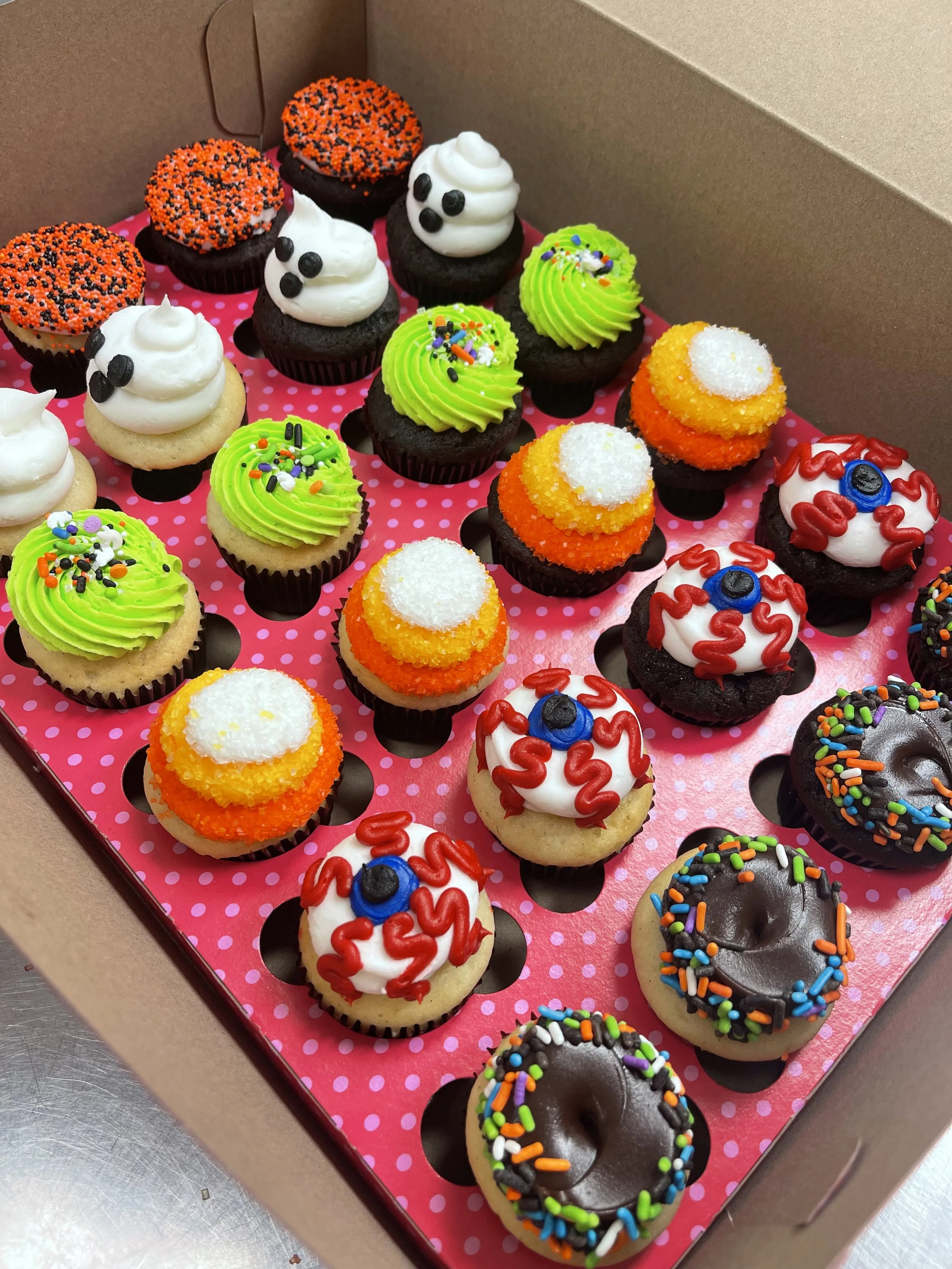 A box of decorated cupcakes with Halloween themes, including ghost faces, eyeballs, and colorful sprinkles.