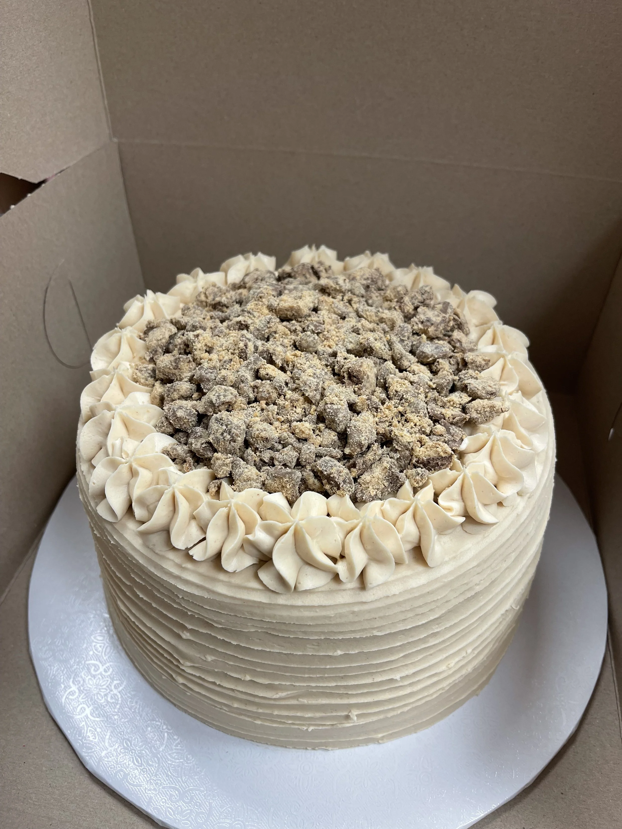 A round cake with cream-colored frosting, decorated with piped frosting around the top edge, topped with crumbled cookies or nuts, inside a cardboard box.