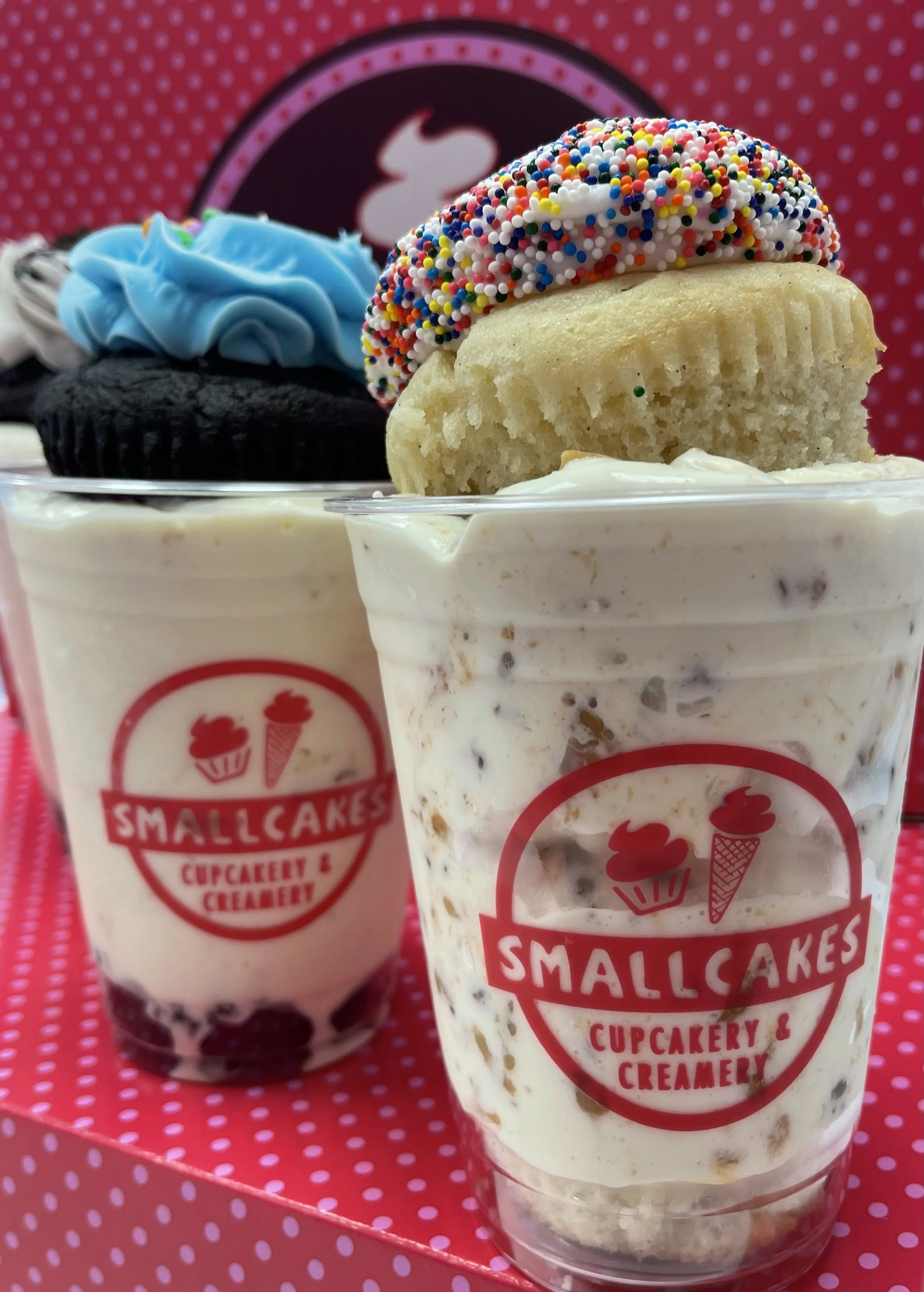 Two cups of ice cream or milkshake from Smallcakes Cupcakery & Creamery with cupcakes on top, one with black frosting and blue icing, and the other with vanilla frosting and colorful sprinkles.