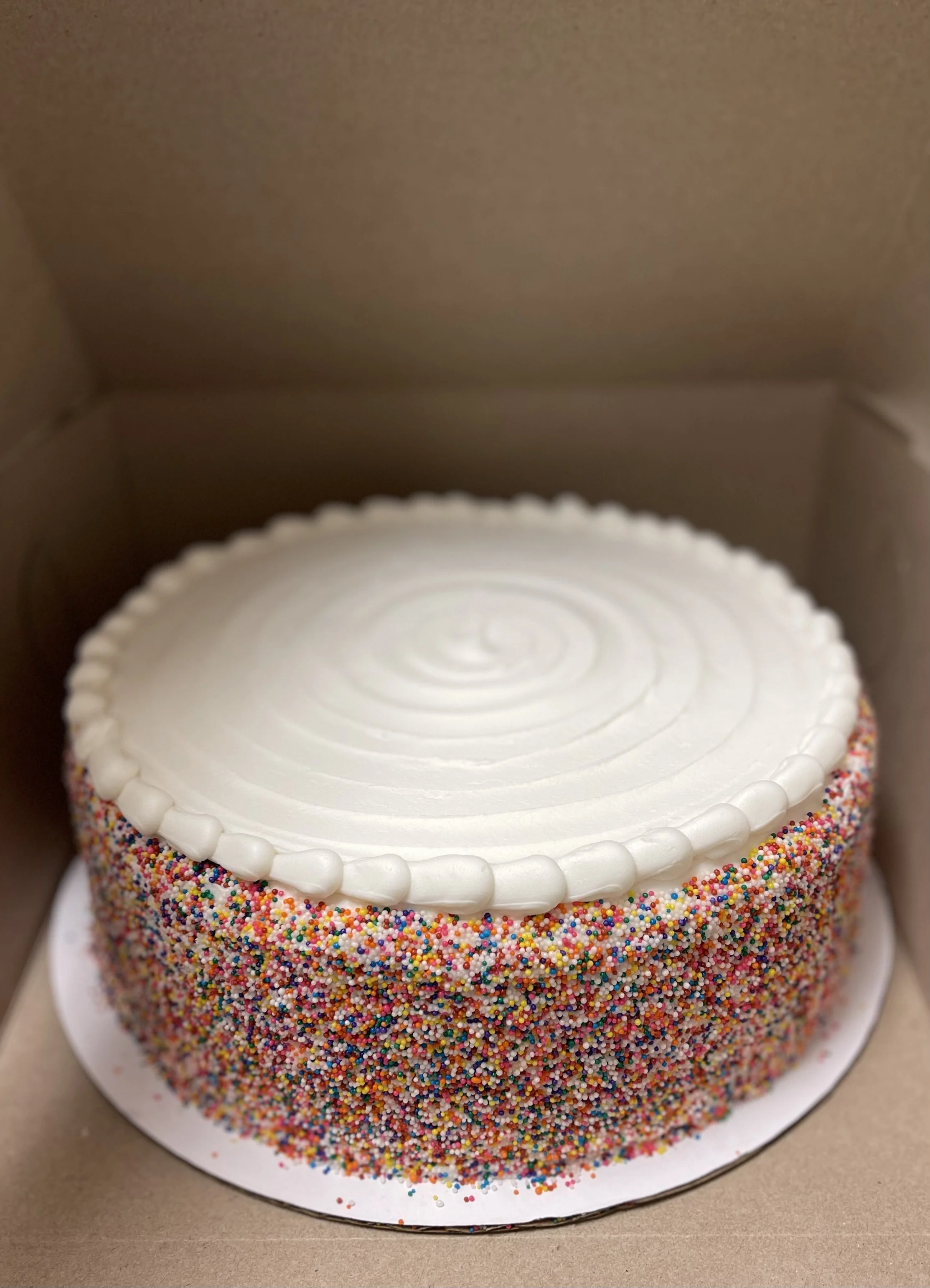 A round cake decorated with multicolored rainbow sprinkles around the sides, topped with white frosting and a border of small white frosting dots, inside a cardboard box.