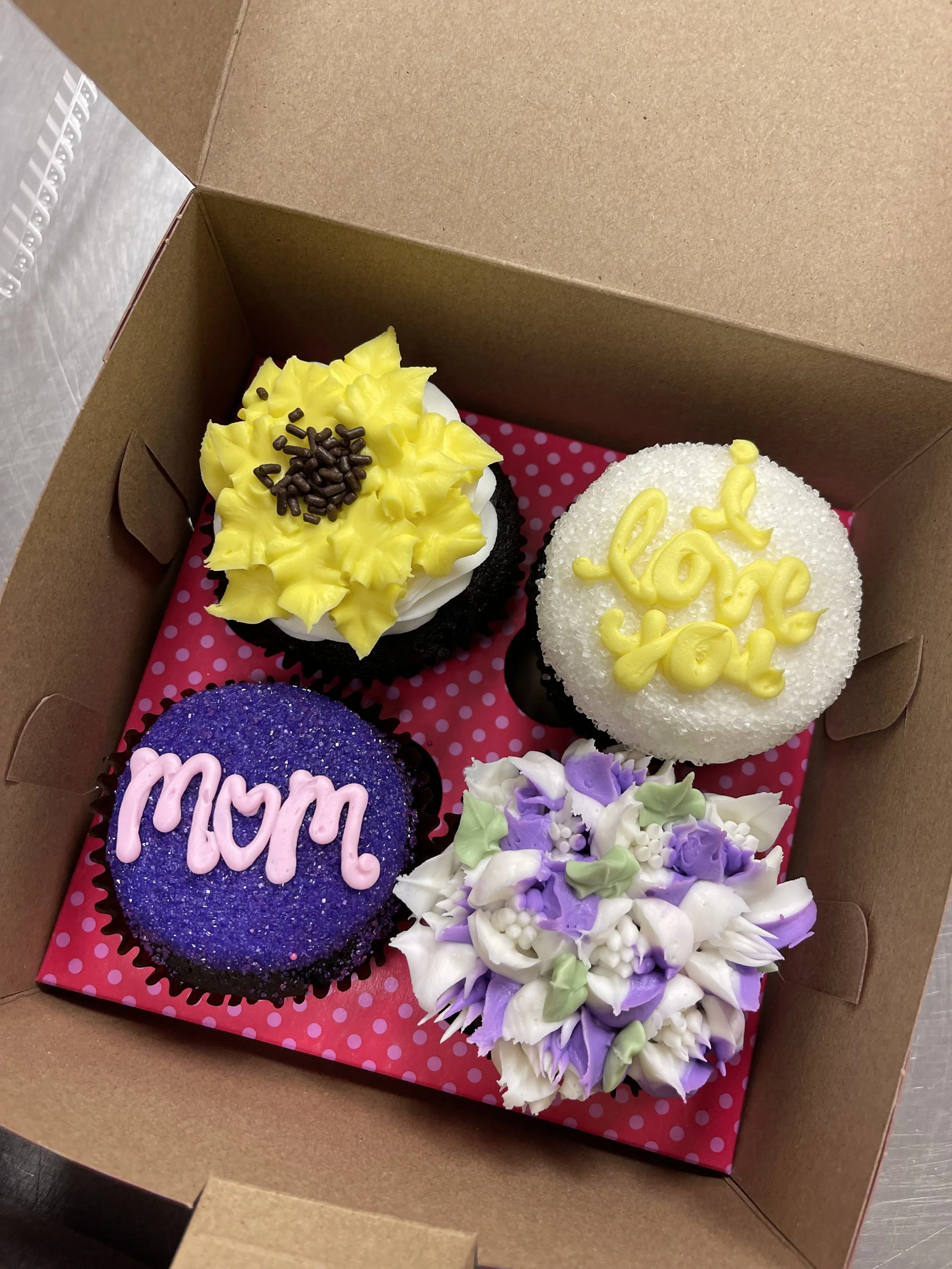 Box of four decorated cupcakes with colorful toppings, arranged on a pink polka-dotted base inside a brown cardboard box.