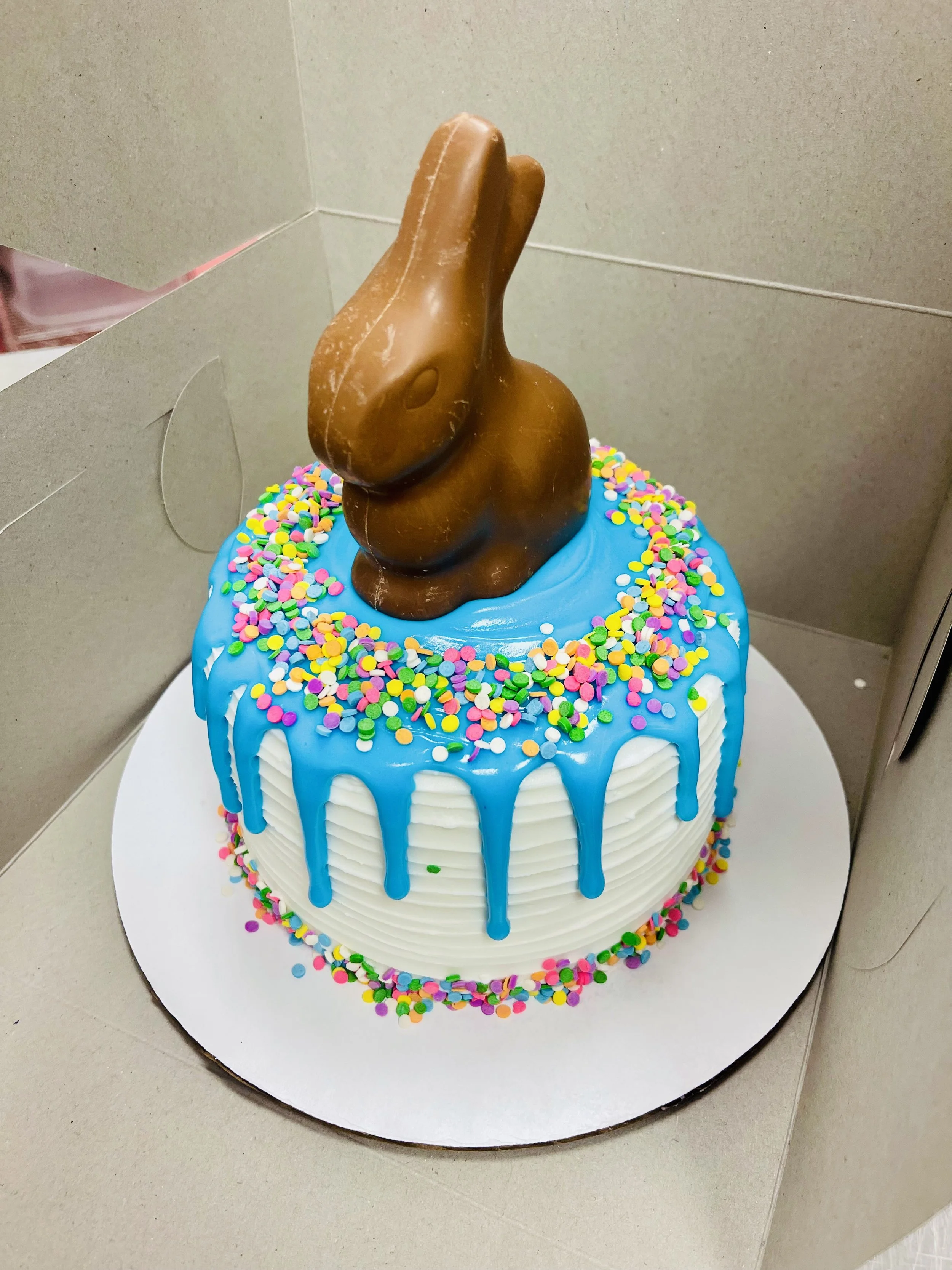 A decorated birthday cake with white frosting, blue drip icing, colorful sprinkles on top and around the base, and a chocolate bunny on top.
