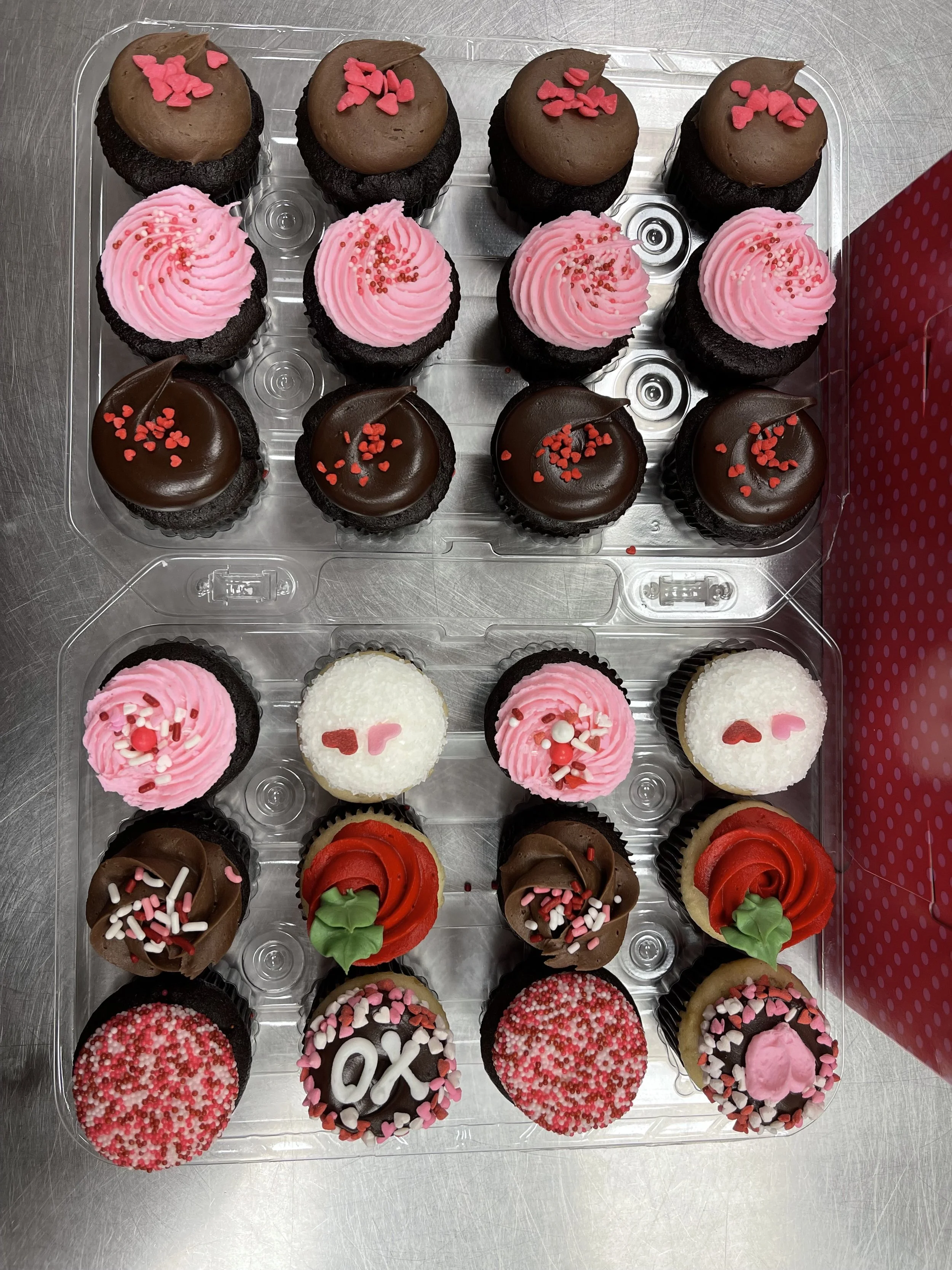 A clear plastic tray holding 20 decorated cupcakes arranged in four rows, each with different frosting colors and sprinkles, including pink, white, red, and chocolate toppings.