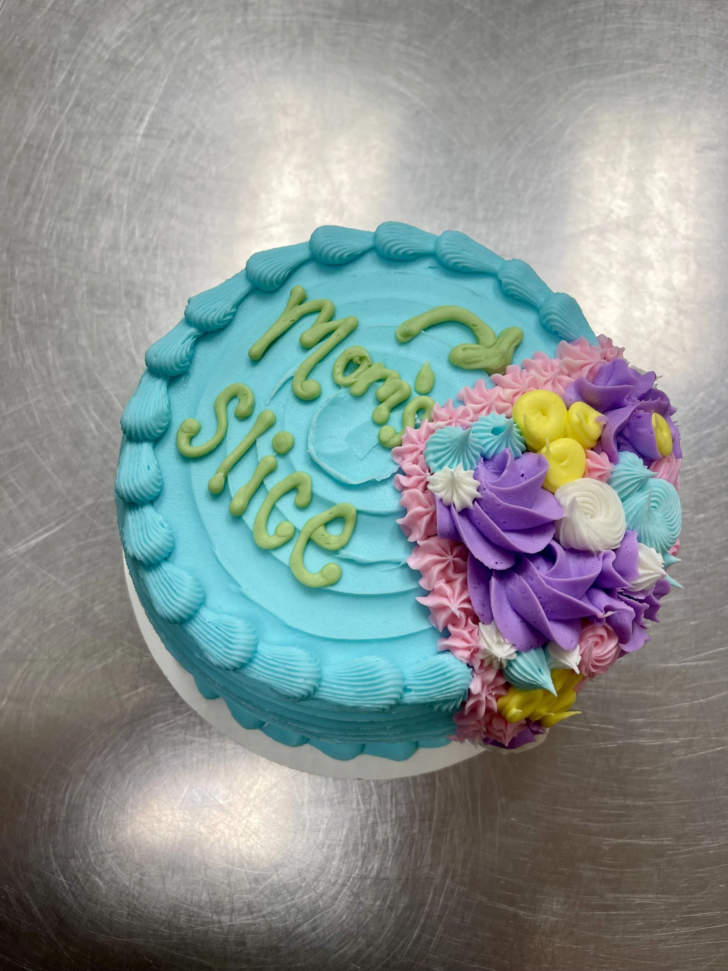 A round birthday cake with blue icing, decorated with pink, purple, white, yellow, and blue frosting flowers, and the words '2 Manic Slice' written in green on top.