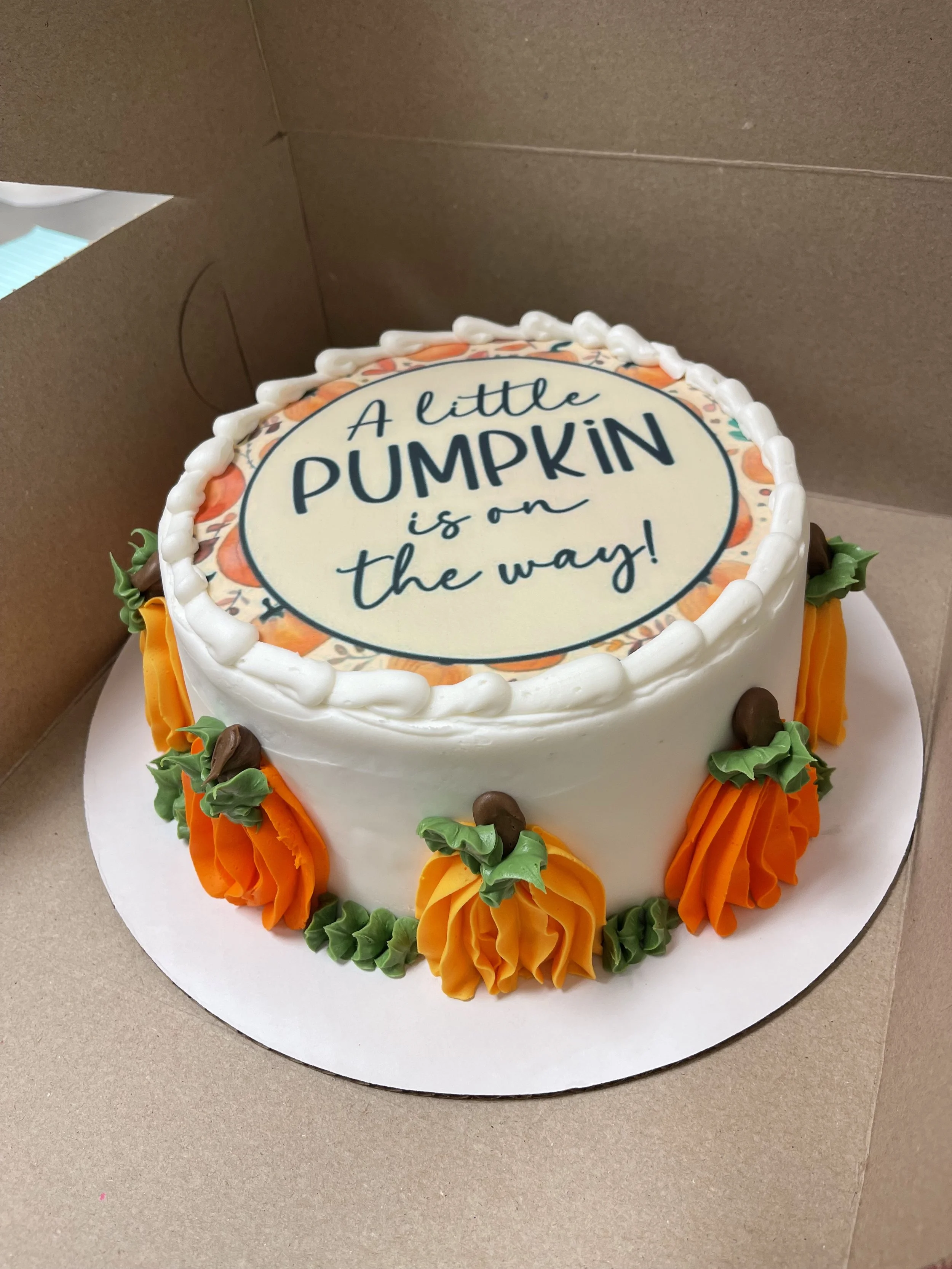 A round birthday cake with white frosting, decorated with orange and green icing flowers, and a top edible decoration with the message: 'A little PUMPKIN is on the way!'