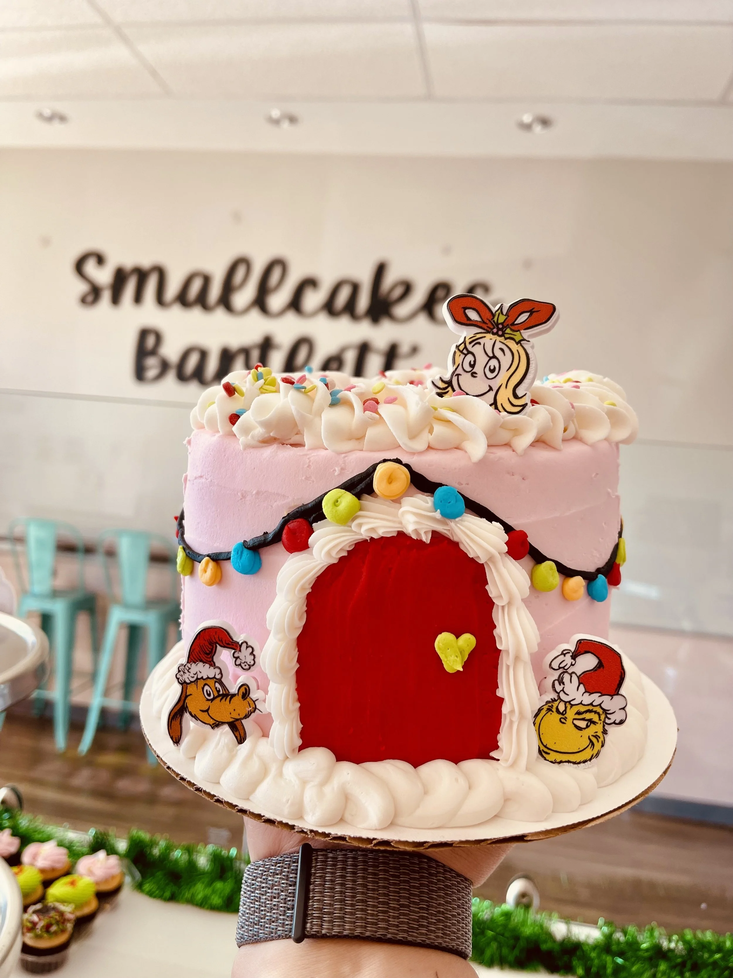 Pink frosted cake decorated with Christmas characters and lights, resembling a house with a door, topped with a cartoon girl and colorful sprinkles, in front of a sign that reads 'smallcakes Bakery'.