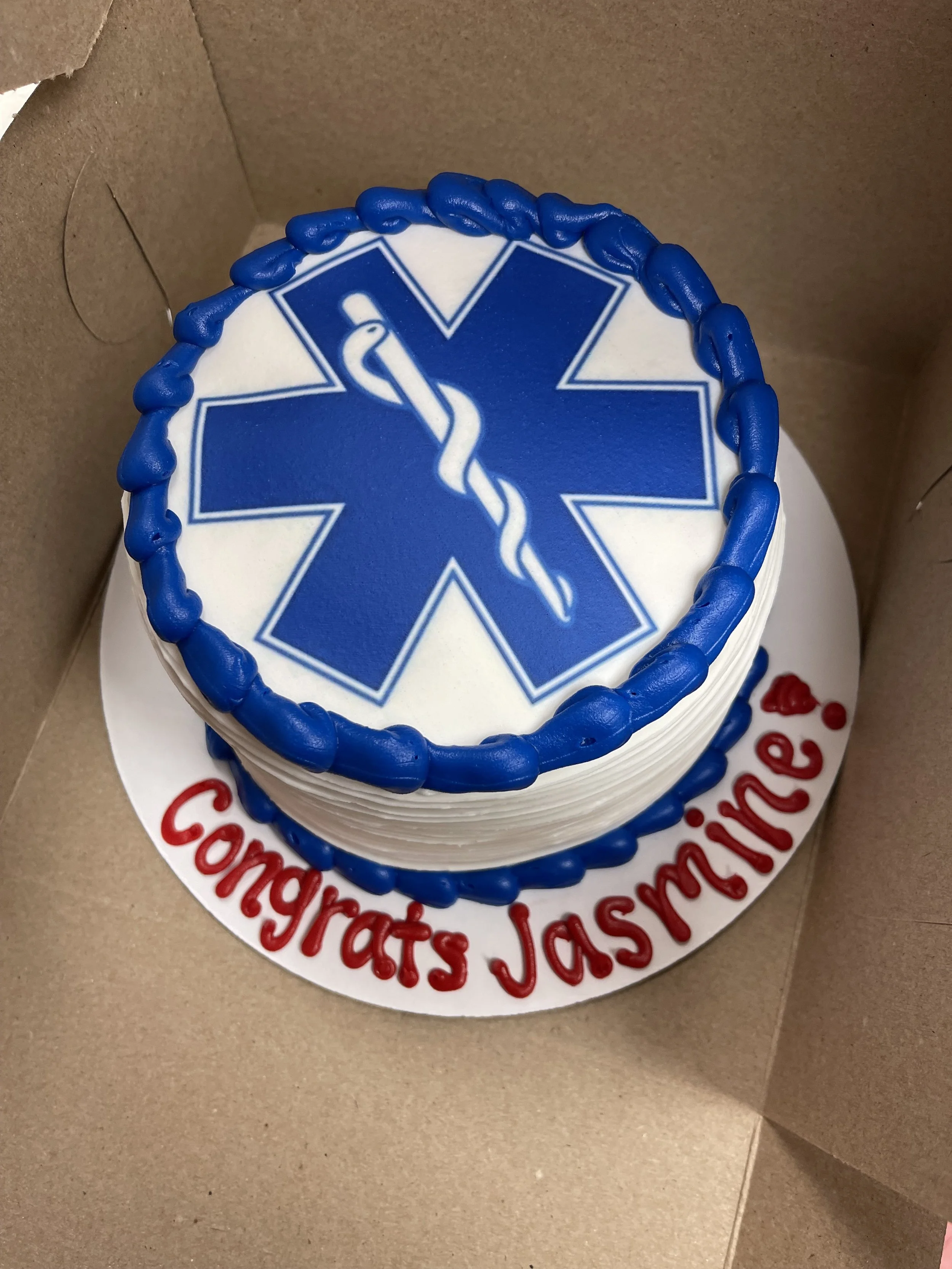 A round cake with white frosting, decorated with blue piping and a blue medical symbol on top. The cake has the words "Congrats Jasminee!" written in red letters on a white base.