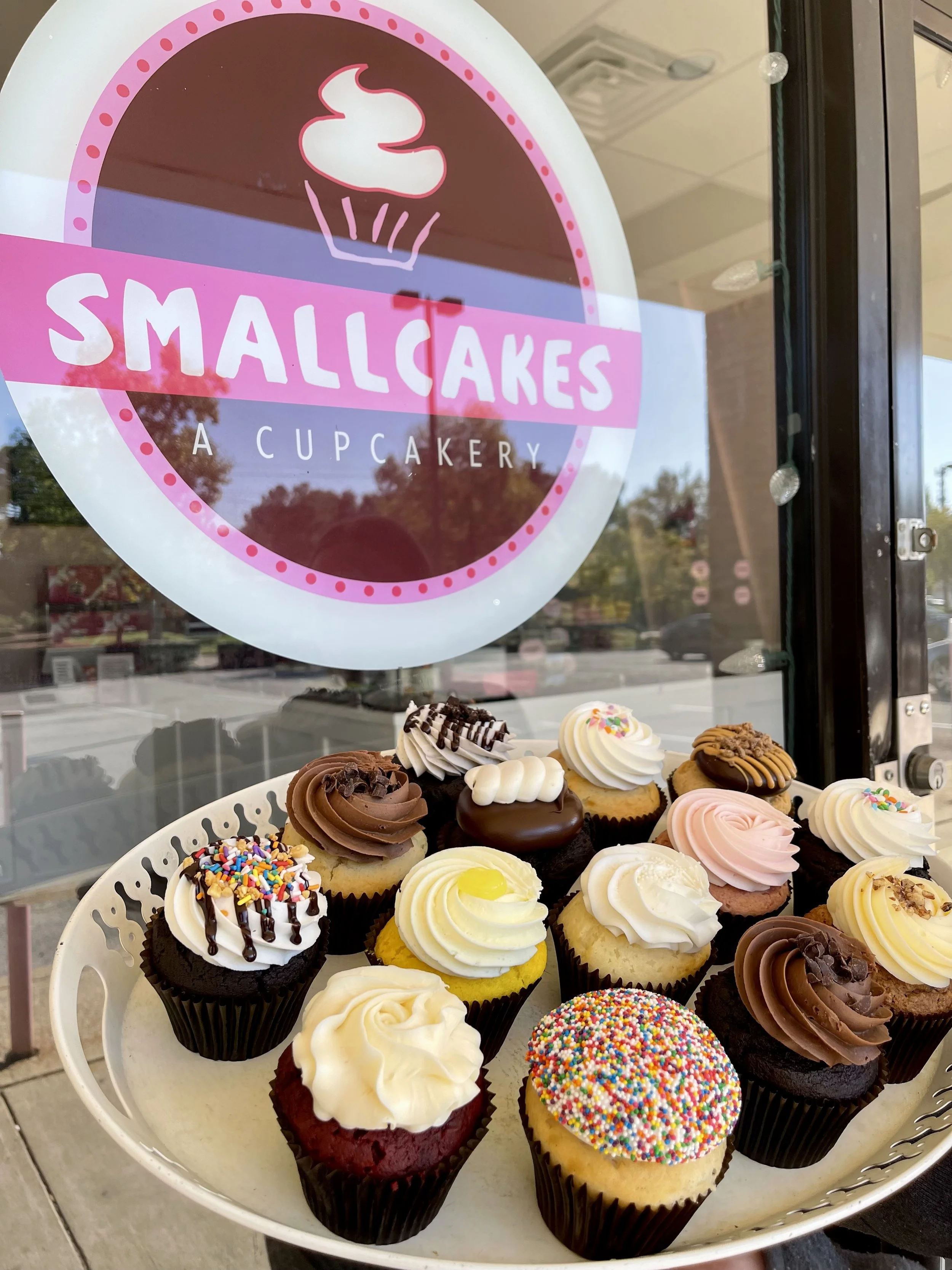 A tray of assorted small cupcakes with various frosting and toppings displayed in front of a bakery window sign that reads 'Smallcakes, A Cupcakery'.